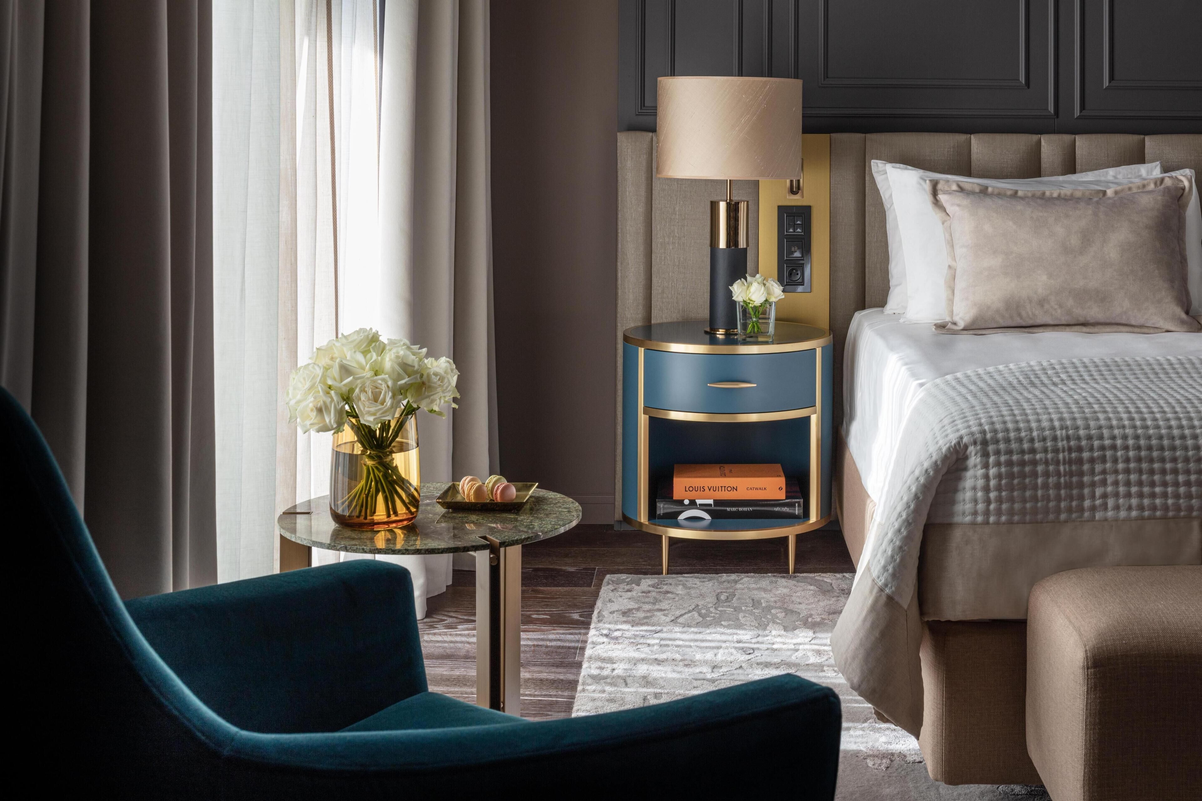 A hotel room with a bed, a nightstand with a lamp, a vase with flowers, and a chair.