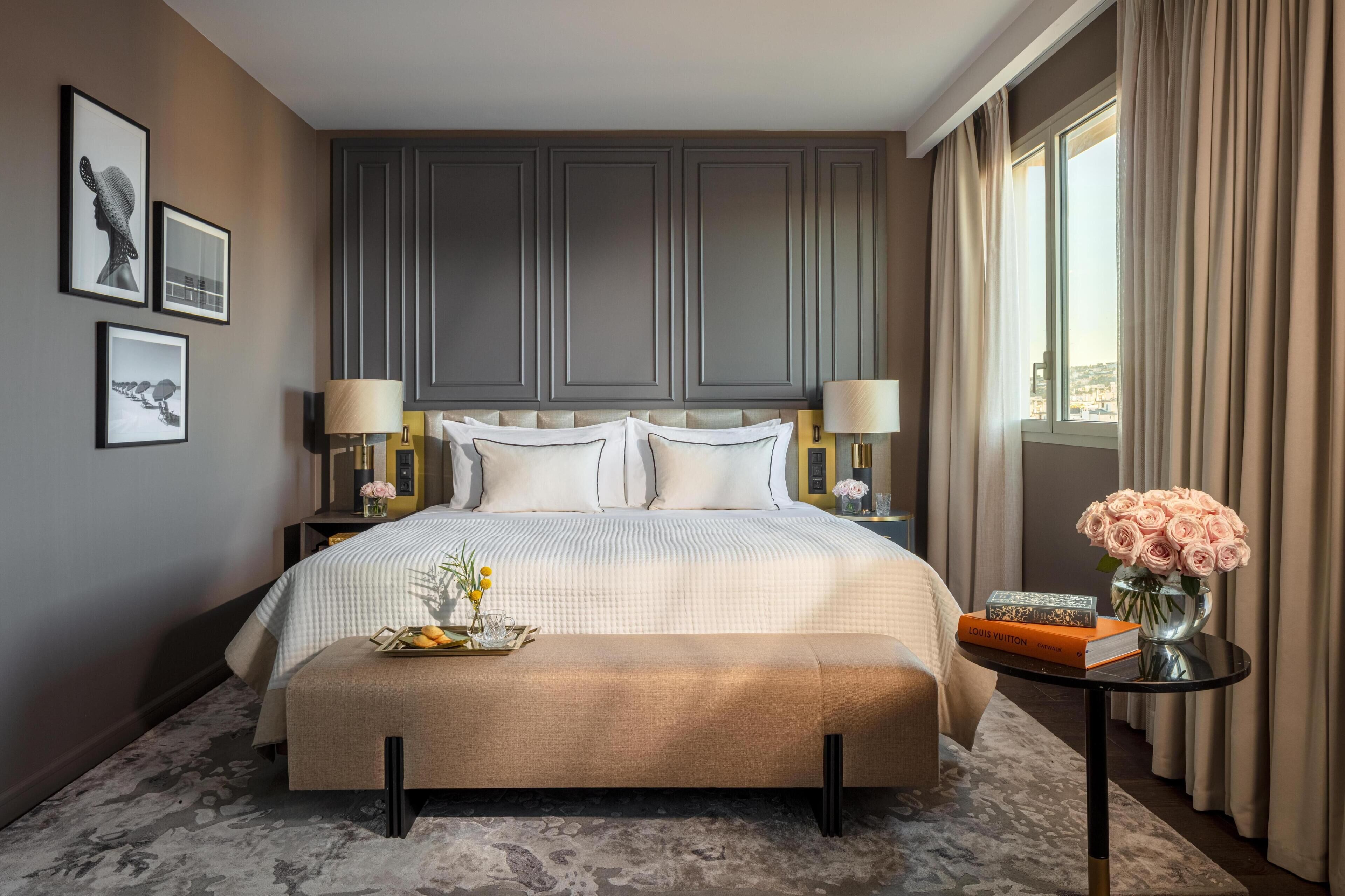 Habitación de hotel con una cama grande, lámparas de noche, una mesita con un jarrón de flores y cuadros en la pared.