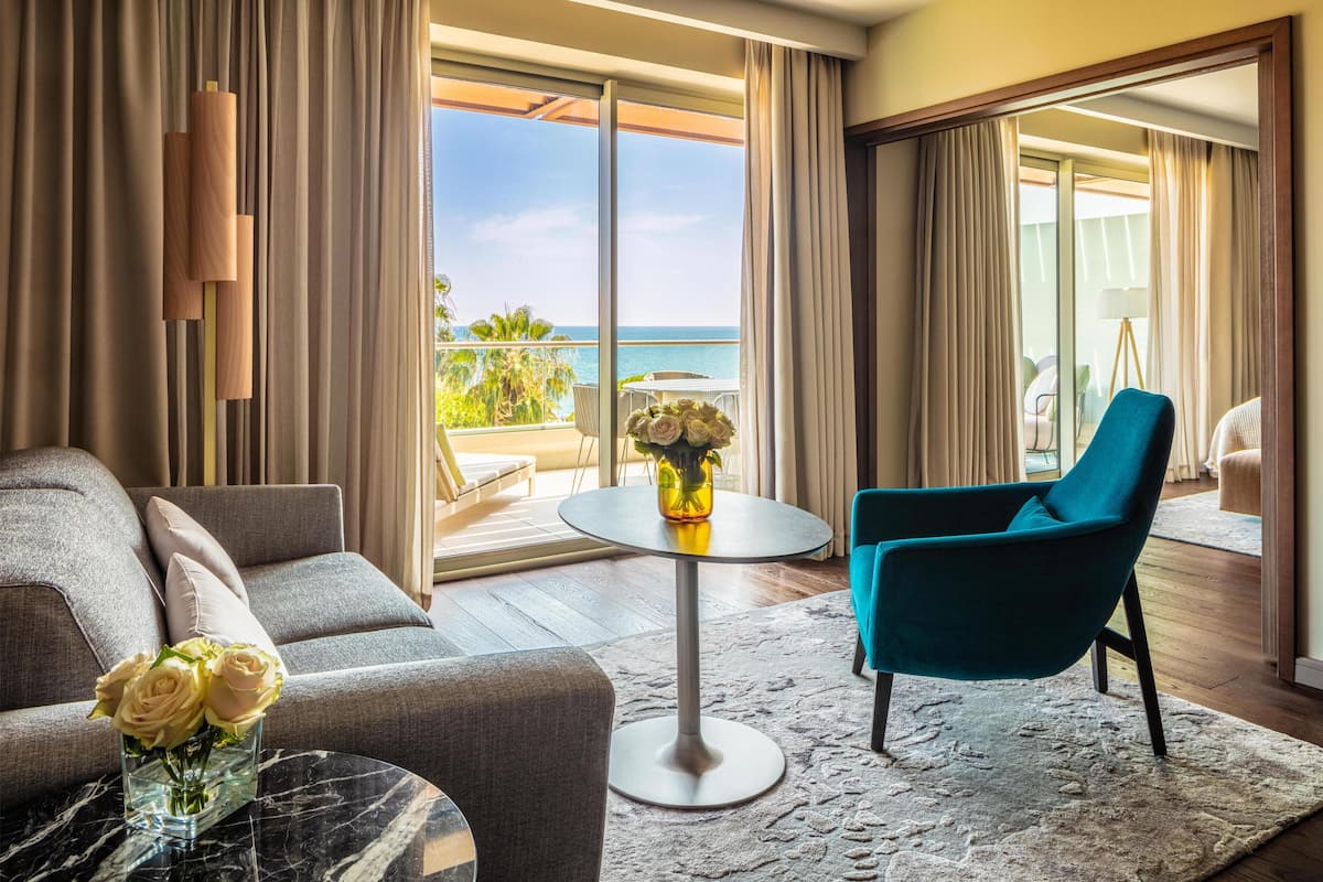 A modern hotel room with a balcony, a sofa, a chair, a small table, and a vase with flowers.