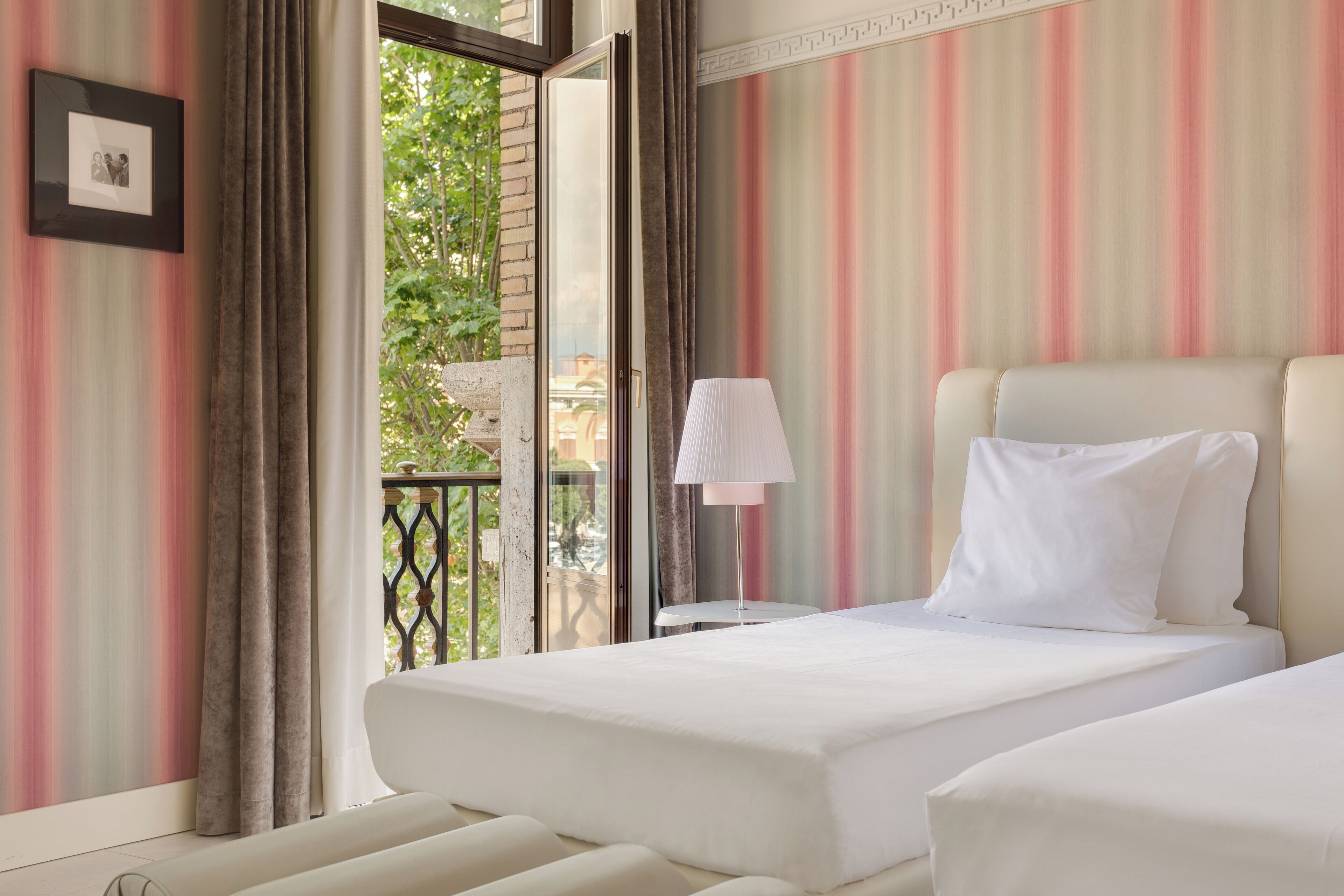 A hotel room with a bed, a bedside lamp, a picture frame, and a view of greenery outside.