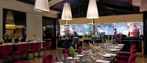 A restaurant with a long dining table set for a meal, red chairs, and overhead pendant lighting.
