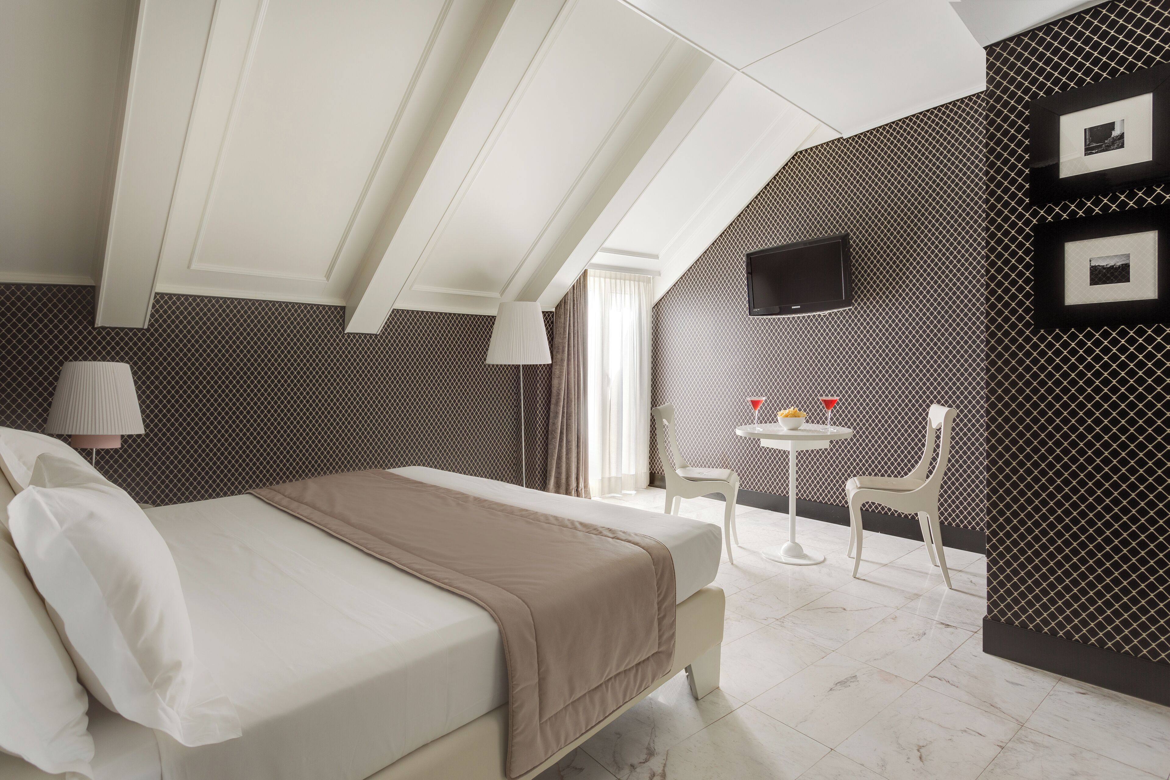 A modern hotel room with a bed, a small table with a lamp, and a TV mounted on the wall.