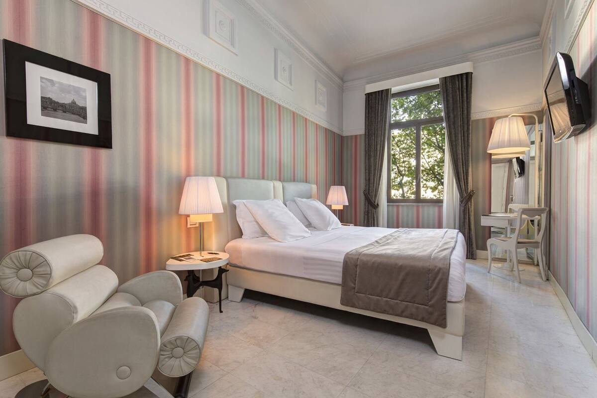 A hotel room with a bed, bedside tables, a chair, a TV, and a window with curtains.
