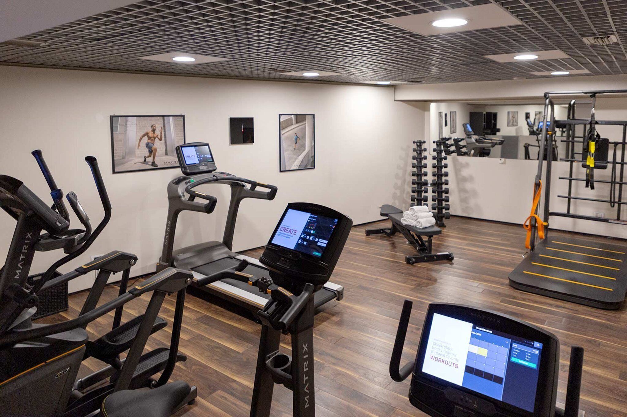 Un gimnasio con cintas de correr, bicicletas estáticas y una máquina de remo. Hay colchonetas de ejercicio y un televisor de pared que muestra videos de entrenamiento.
