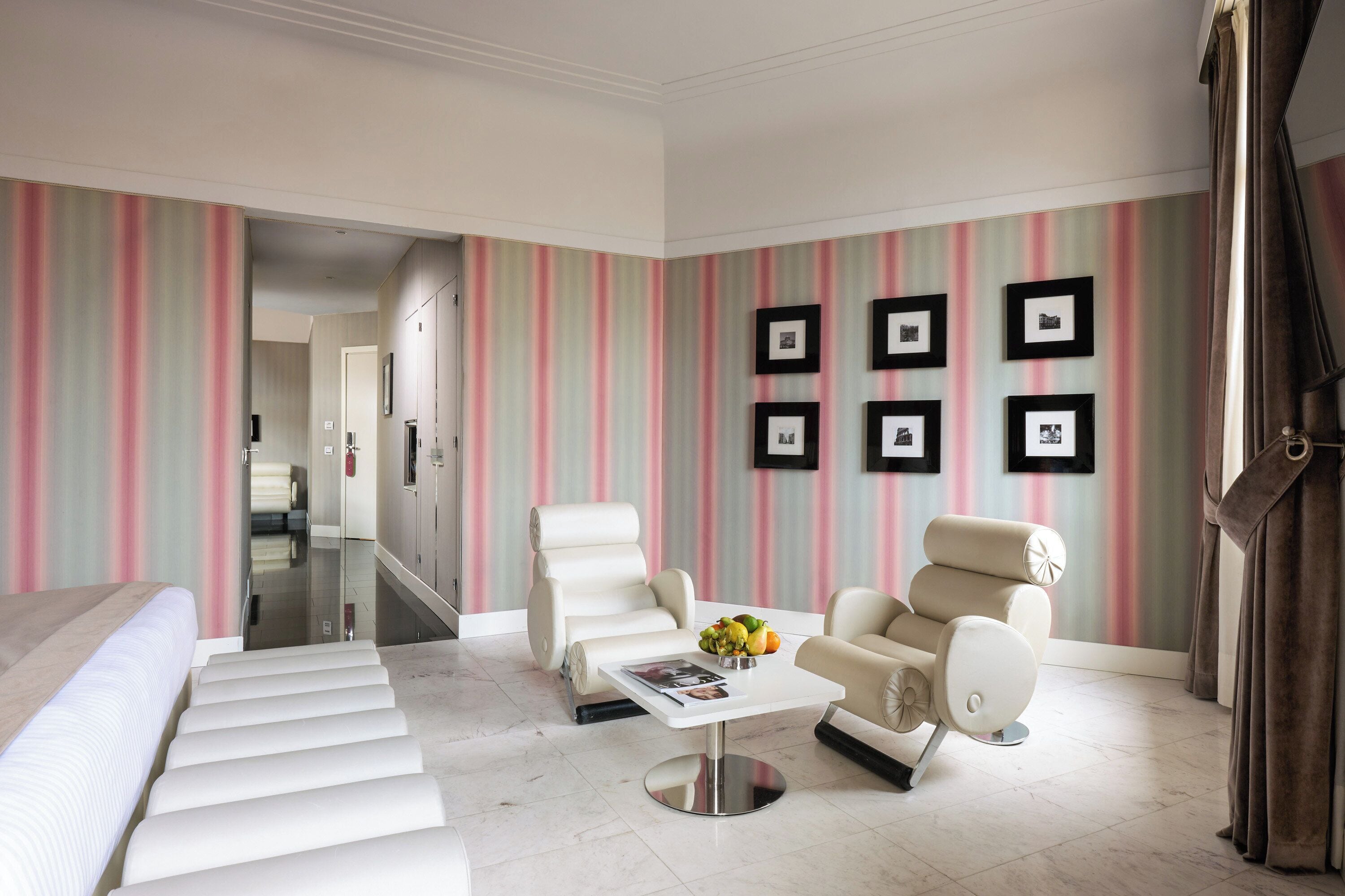 A modern hotel room with a staircase, a white sofa, a beige armchair, a coffee table with fruit, and striped wallpaper.