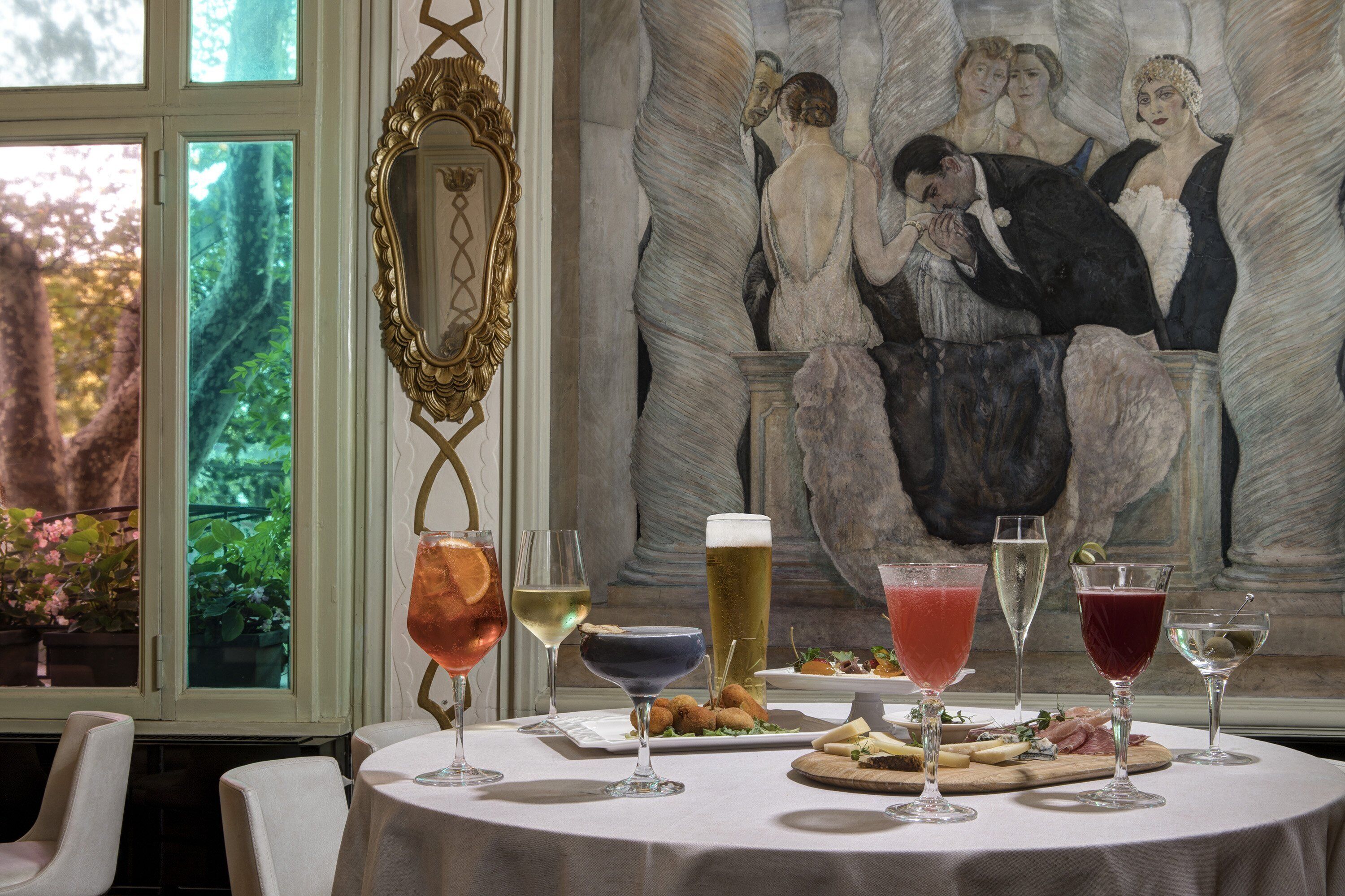 A table set with various drinks and a cheese board, with a painting and a window in the background.