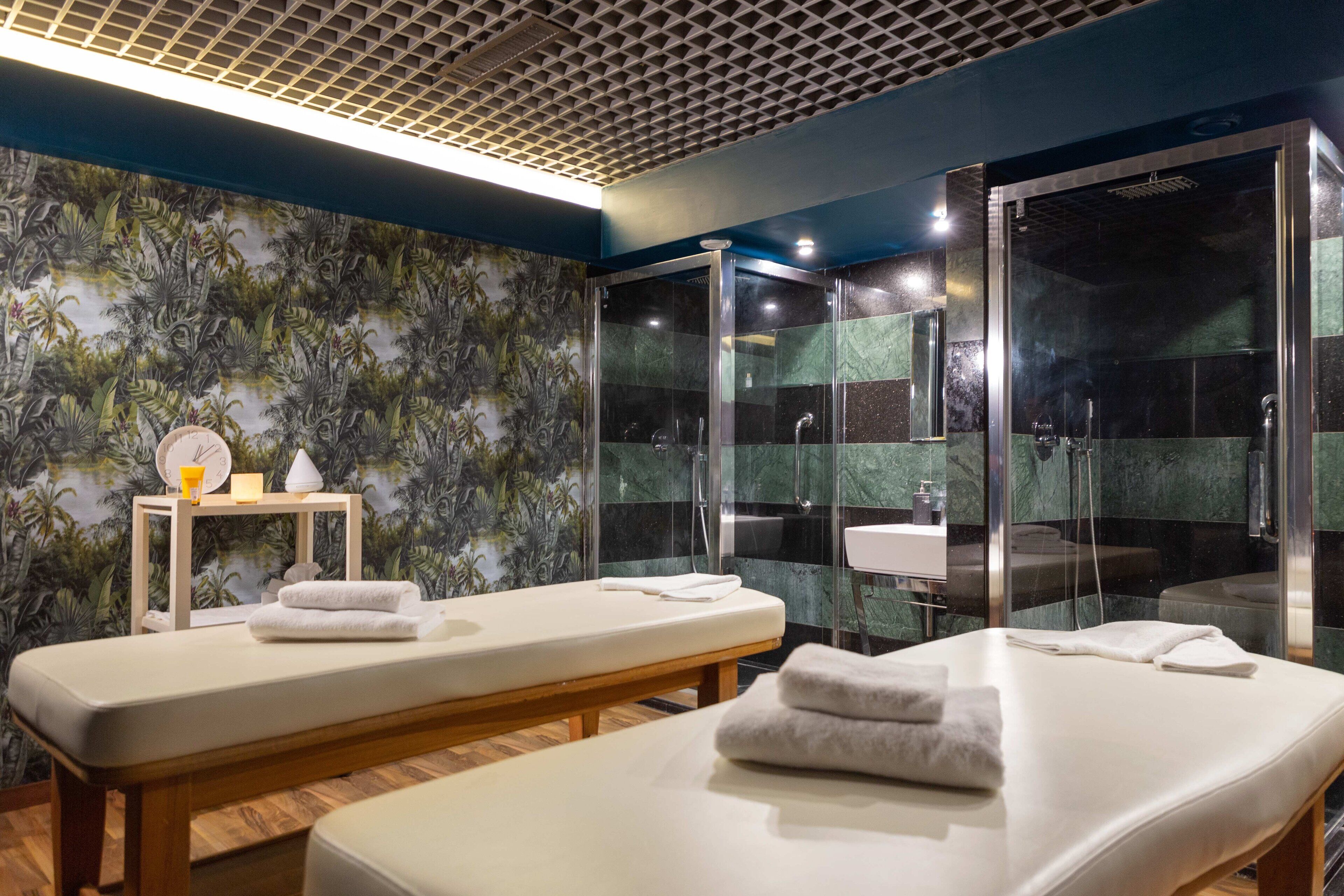 A spa room with two massage tables, a shower area, and a wall with a tropical leaf pattern.