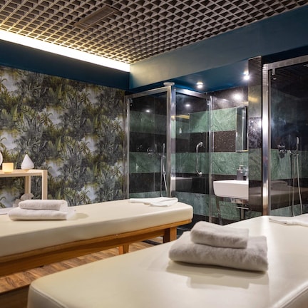 A spa room with two massage tables, a shower area, and a wall with a tropical leaf pattern.