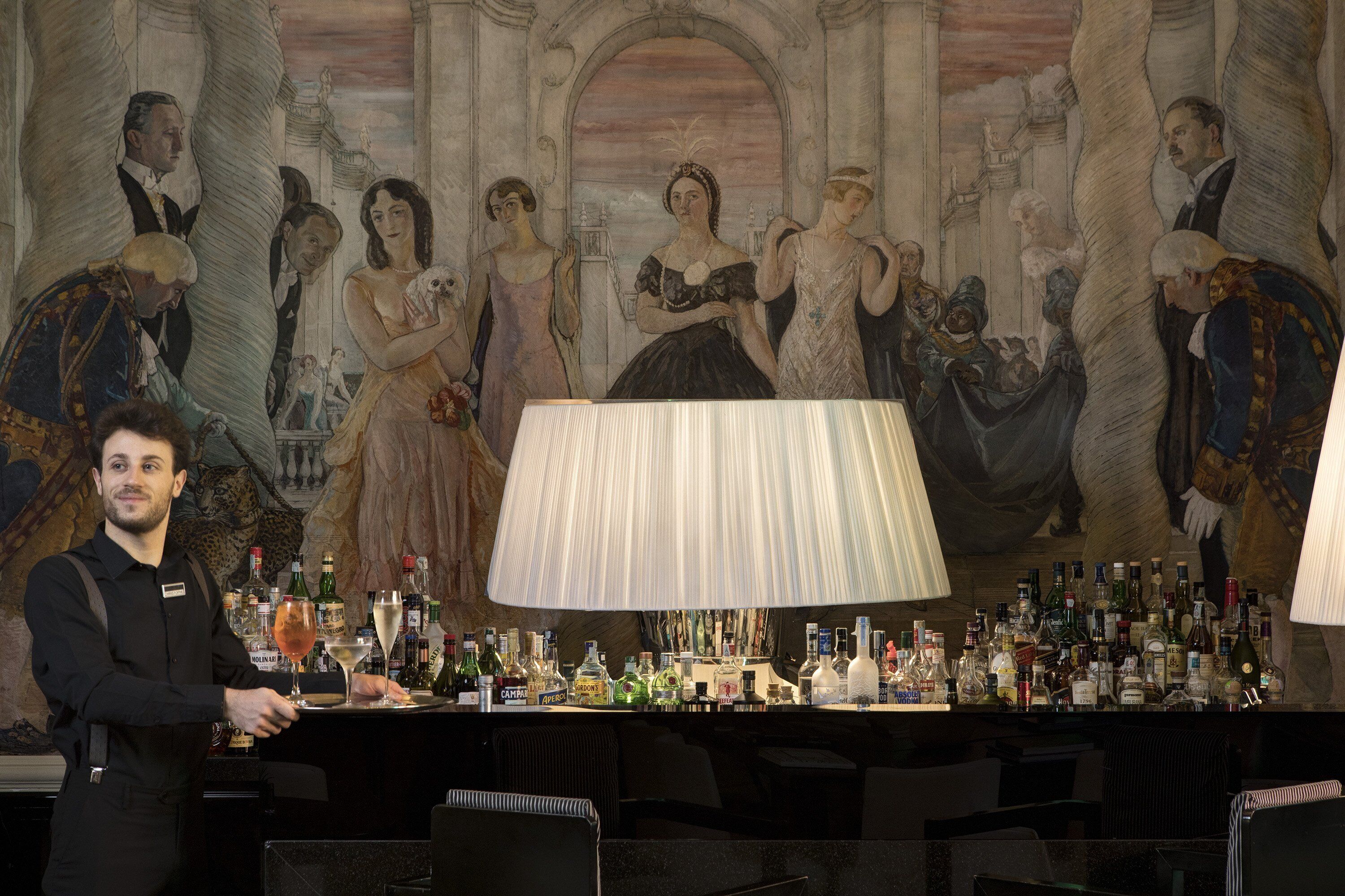 A bar area with a large lamp, a bartender, and a mural with figures in period clothing.