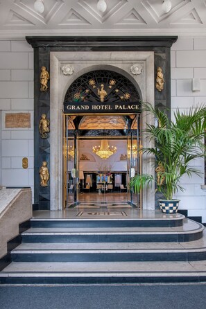 The entrance of Grand Hotel Palace with a grand arched doorway, a chandelier, and a potted plant.