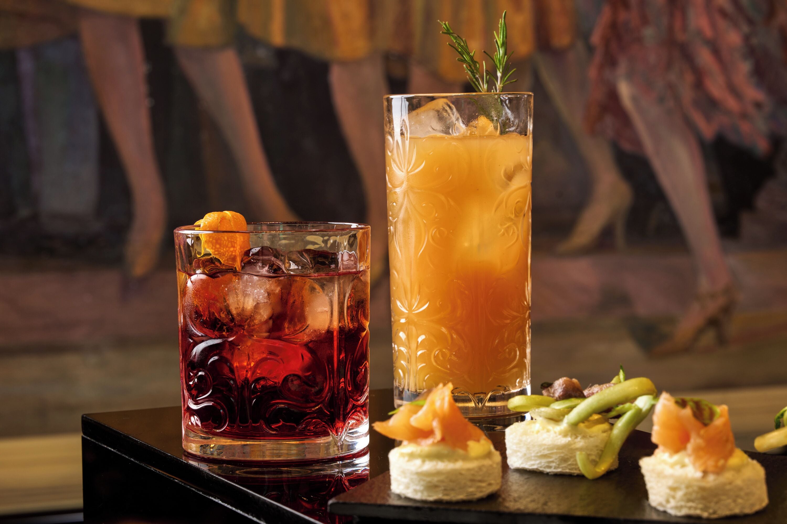 A bar setting with a glass of red beverage, a tall glass of orange drink, and small appetizers on a tray.
