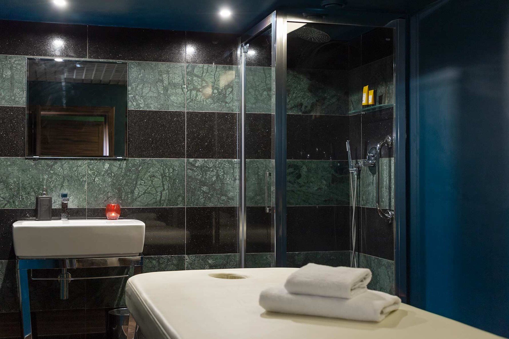 A modern bathroom with a glass shower enclosure, a white sink, and a bed with a white towel.