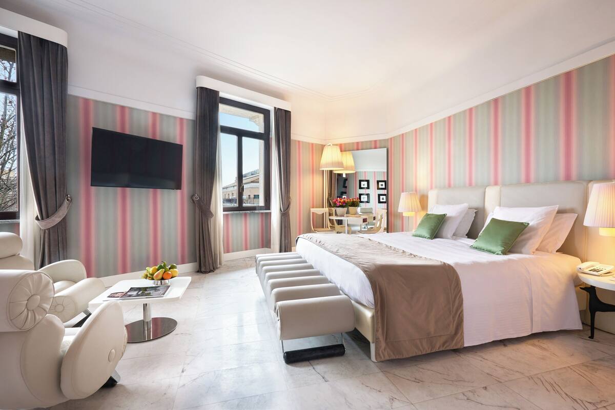 A modern hotel room with a large bed, a seating area with a table, a flat-screen TV, and a view of the city through the window.