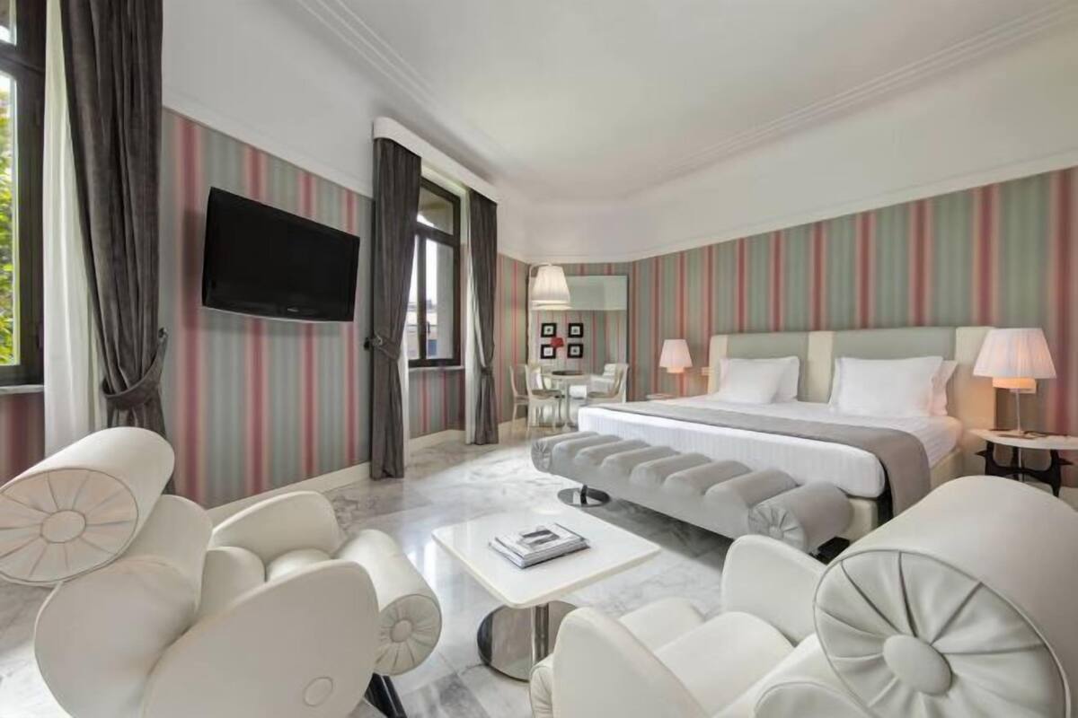 A modern hotel room with a large bed, a flat-screen TV, a dining area, and a view of the outdoors.