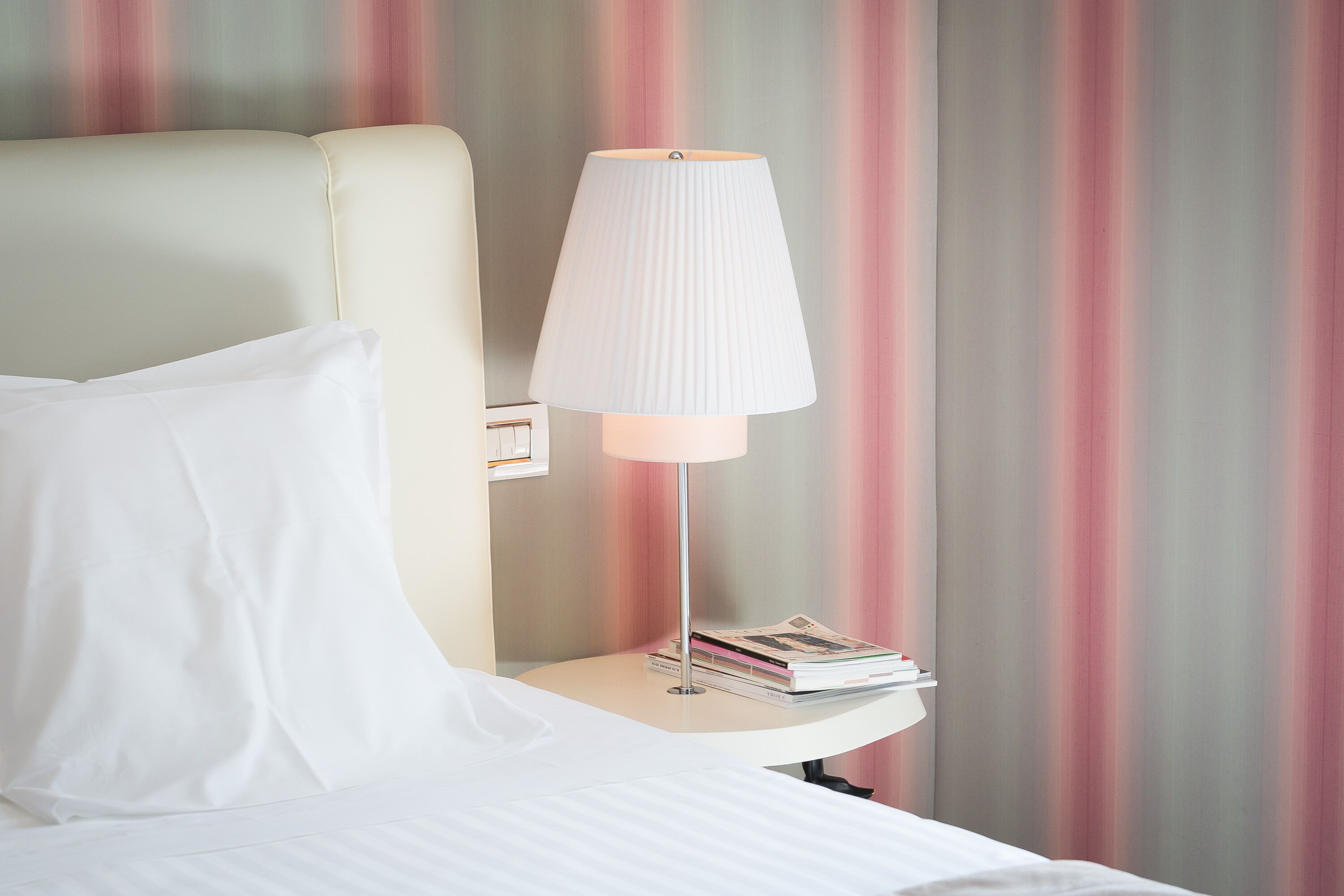 A neatly made bed with white linens, a bedside lamp, and a stack of books on a small table beside the bed.