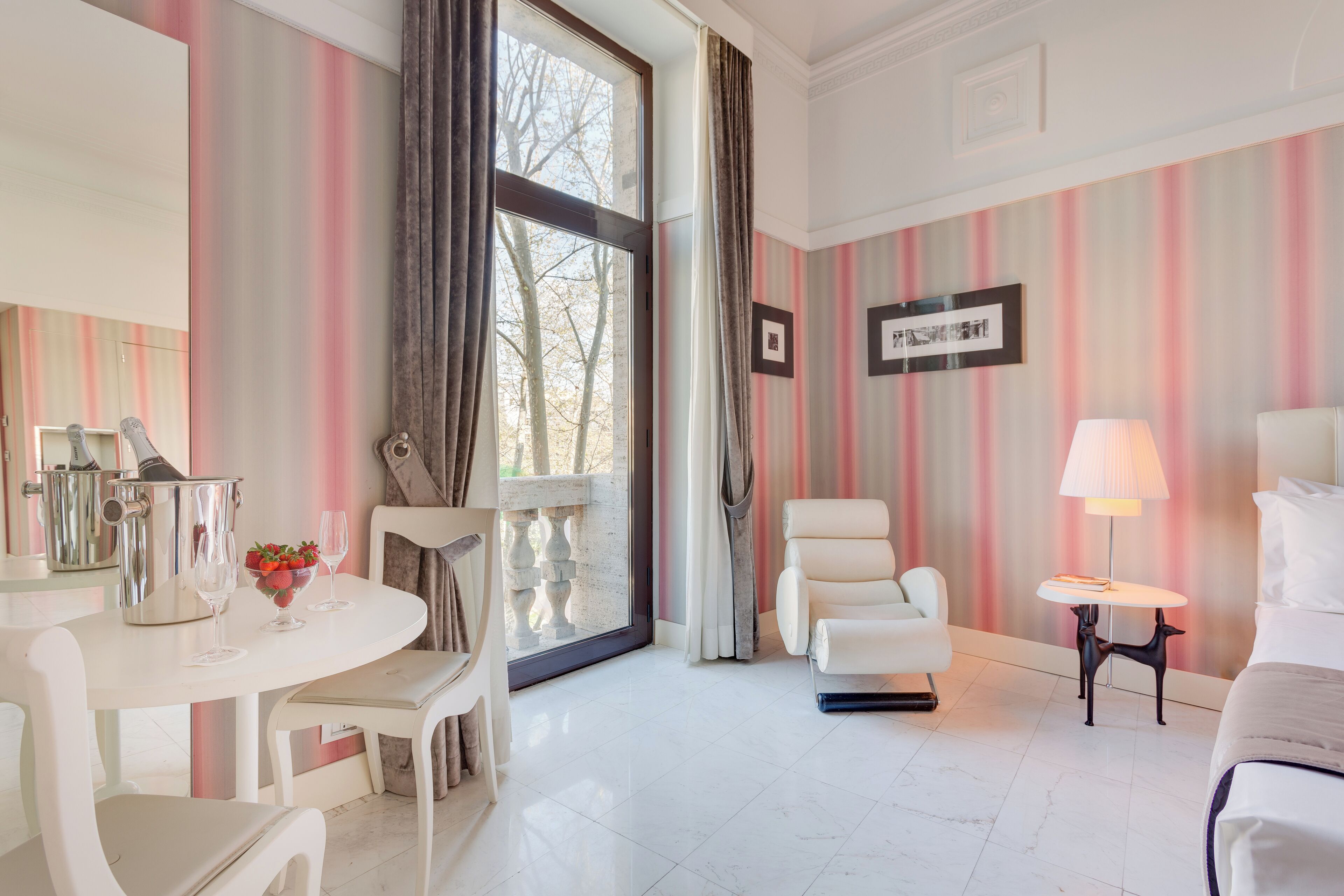 Camera d'albergo con tavolo da pranzo, sedie bianche, una poltrona bianca, un letto, un comodino con una lampada e carta da parati a righe.