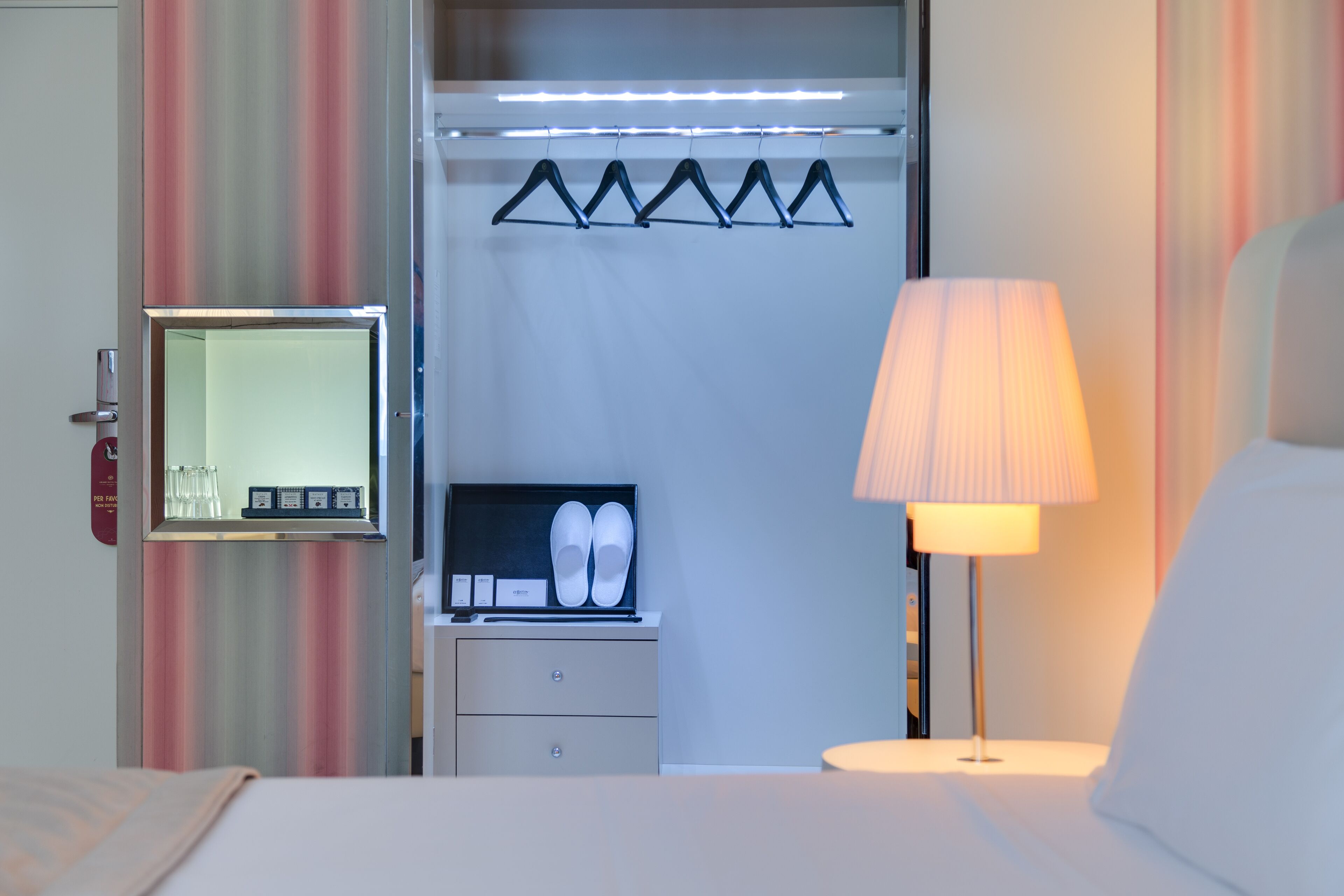 Habitación de hotel con cama, mesita de noche con lámpara, armario con espejo y zapatero.