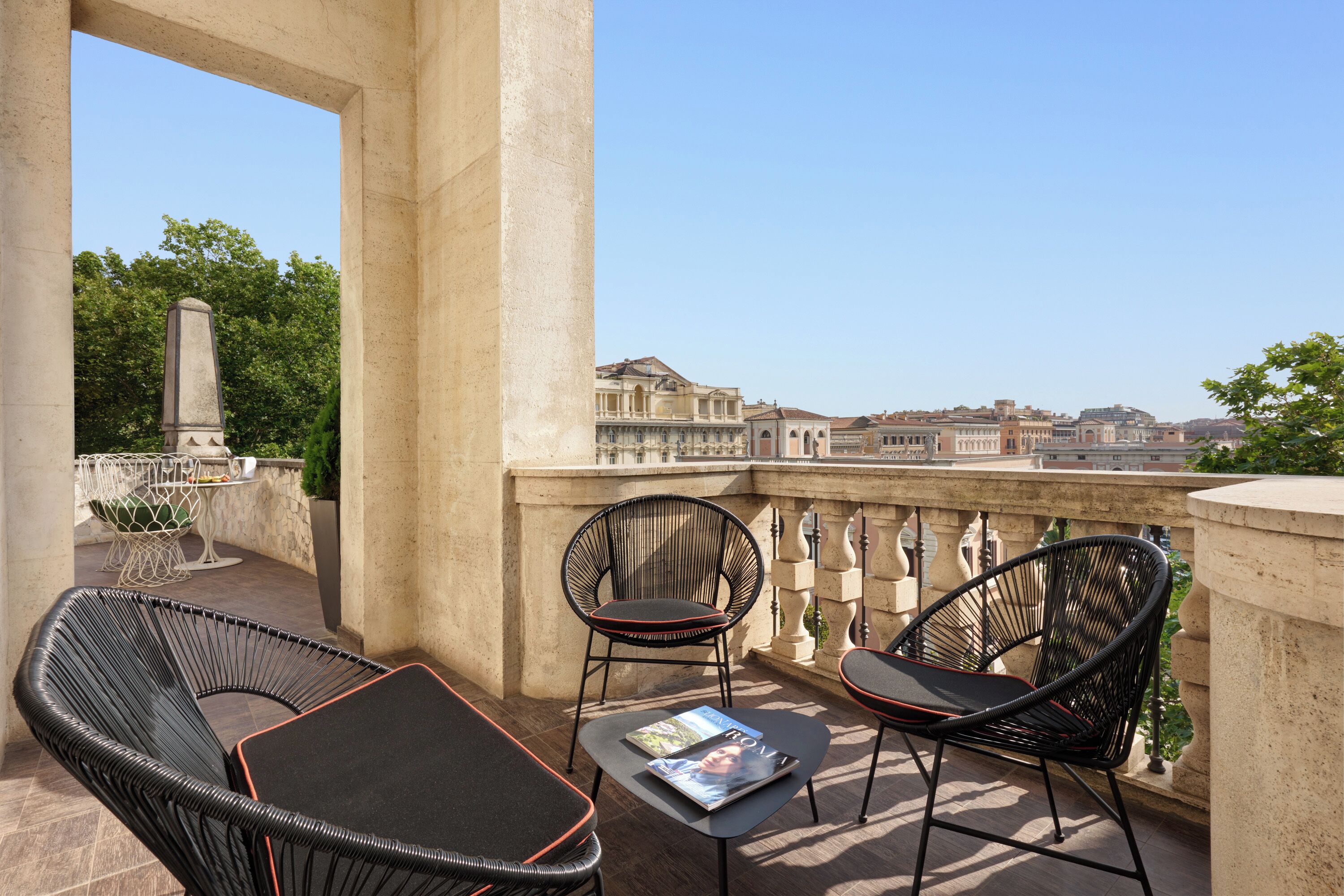 A balcony with wicker chairs and a table, overlooking a cityscape with historic buildings.