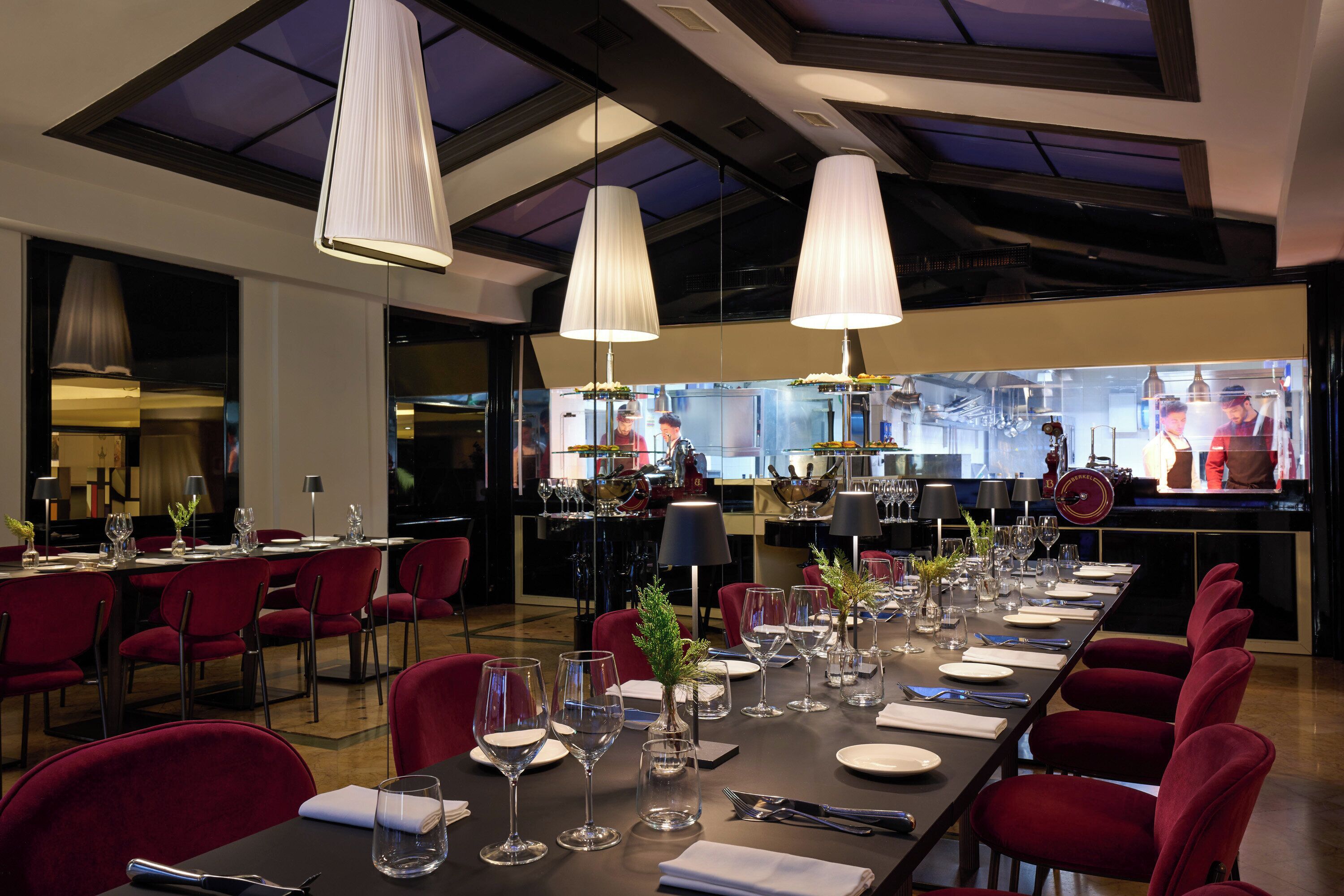 Un restaurant avec une longue table à manger dressée pour le repas, des chaises rouges et des lampes suspendues.