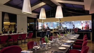 A restaurant with a long dining table set for a meal, red chairs, and hanging pendant lights.