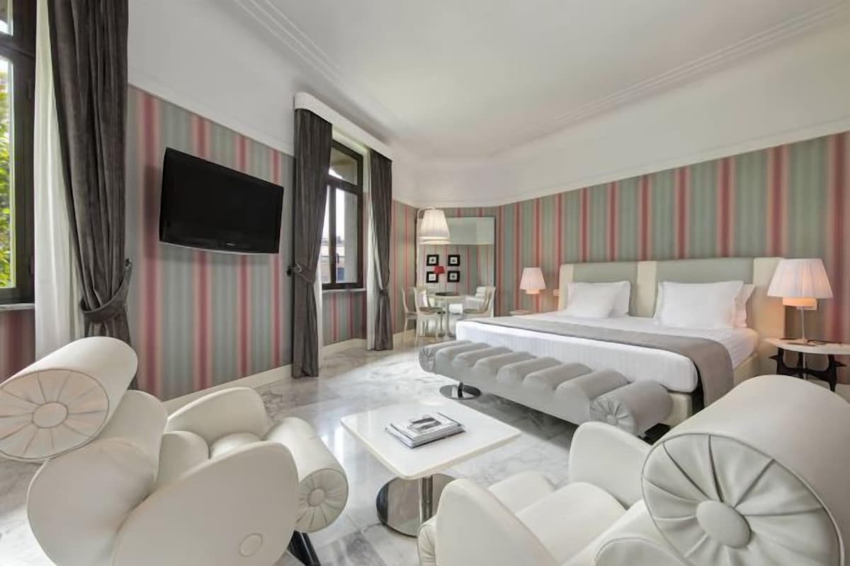 A modern hotel room with a large bed, a flat-screen TV, a dining area, and a view of the outdoors.