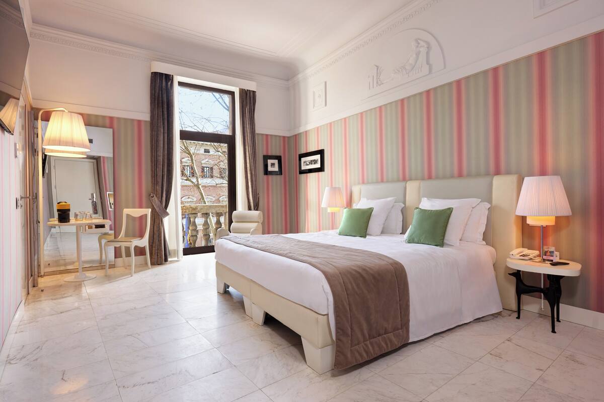 A hotel room with a bed, bedside tables, a chair, a desk, and a view through a window.