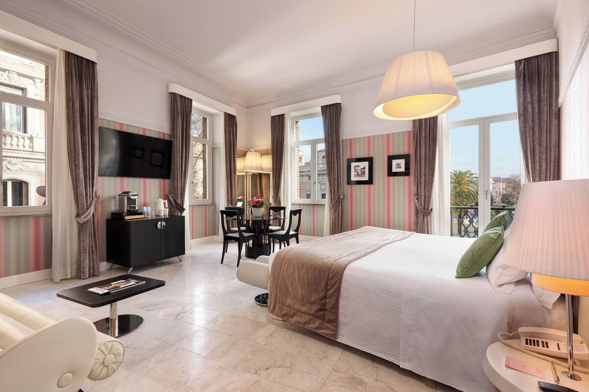 A hotel room with a large bed, a desk with a chair, a television, and a balcony with curtains.