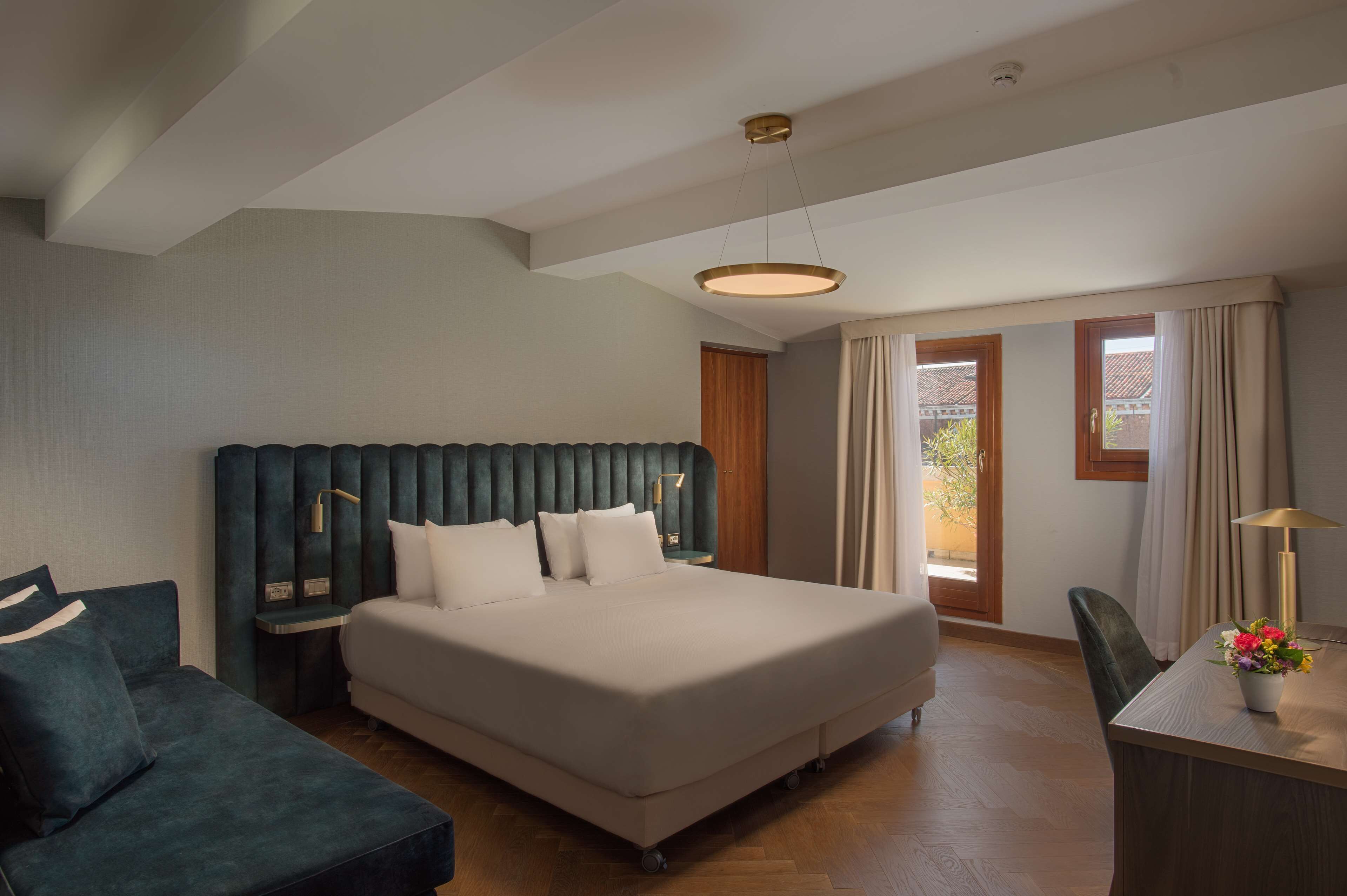 A hotel room with a large bed, a desk, and a chair. There is a window with curtains and a view of the outside.