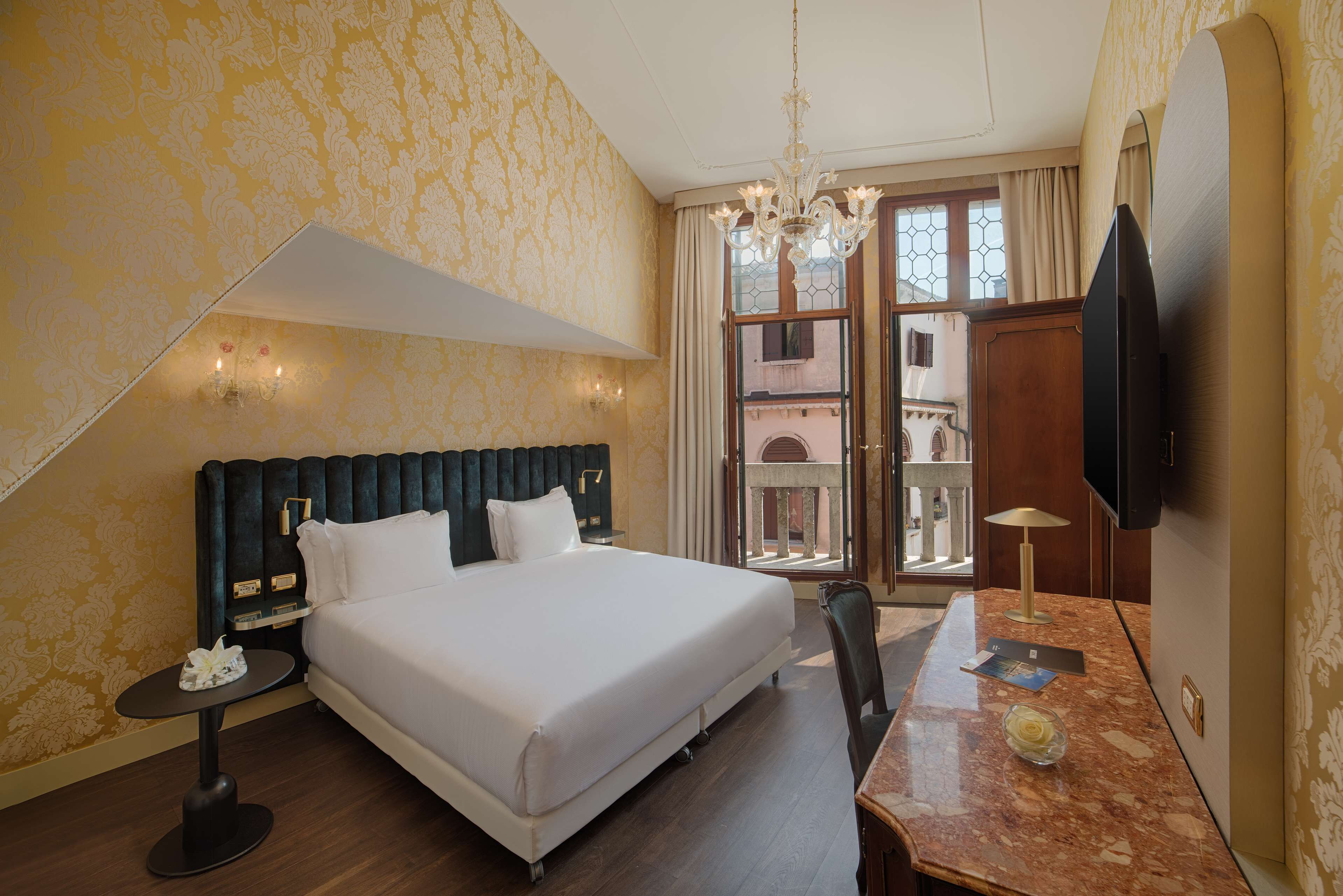 A hotel room with a large bed, a desk with a lamp, a chandelier, and a TV.