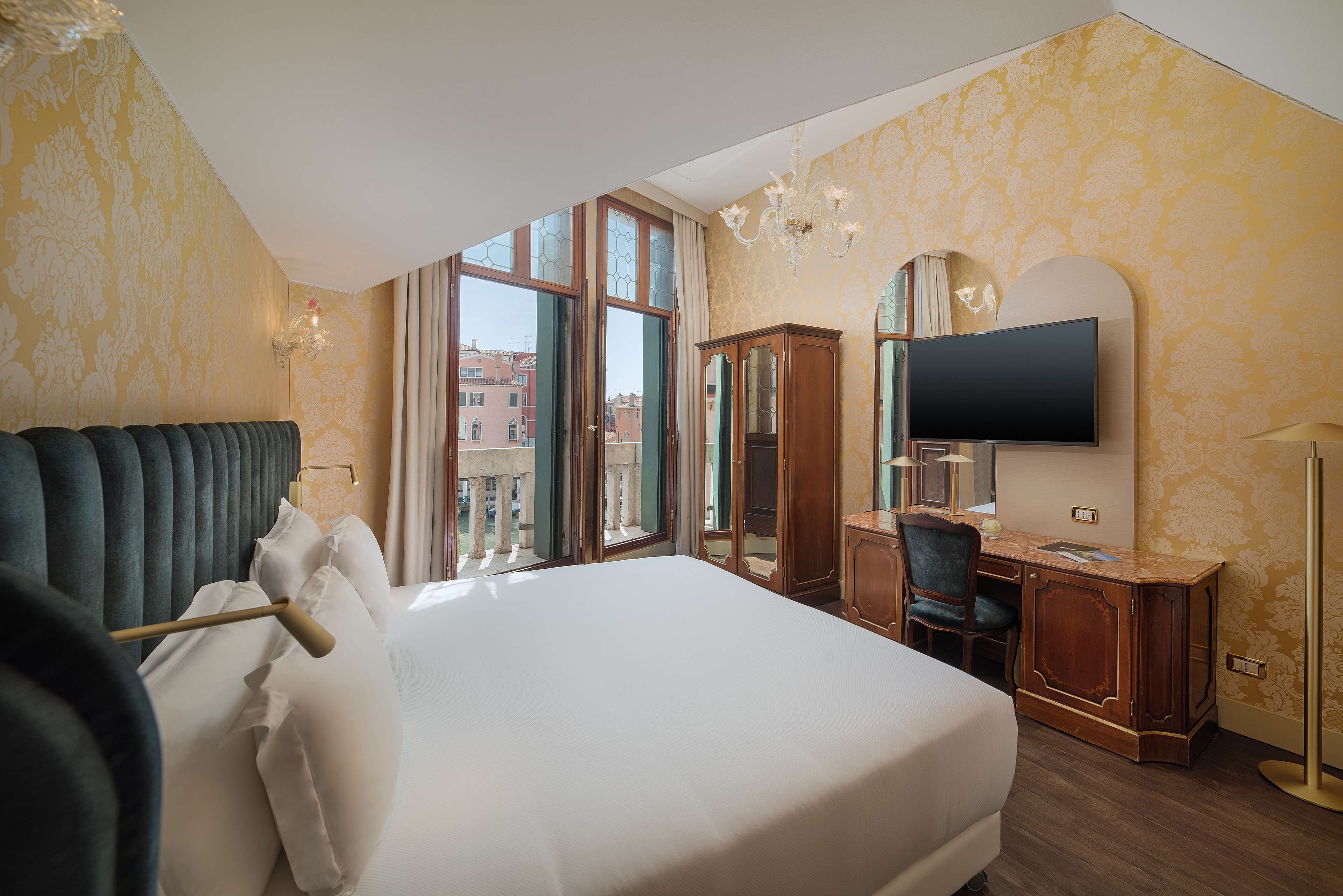 Habitación de hotel con una cama grande, un escritorio, televisión y vistas de la ciudad a través de amplios ventanales.