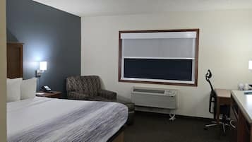 A hotel room with a bed, a desk, a chair, a window with blinds, a heater, and a lamp.