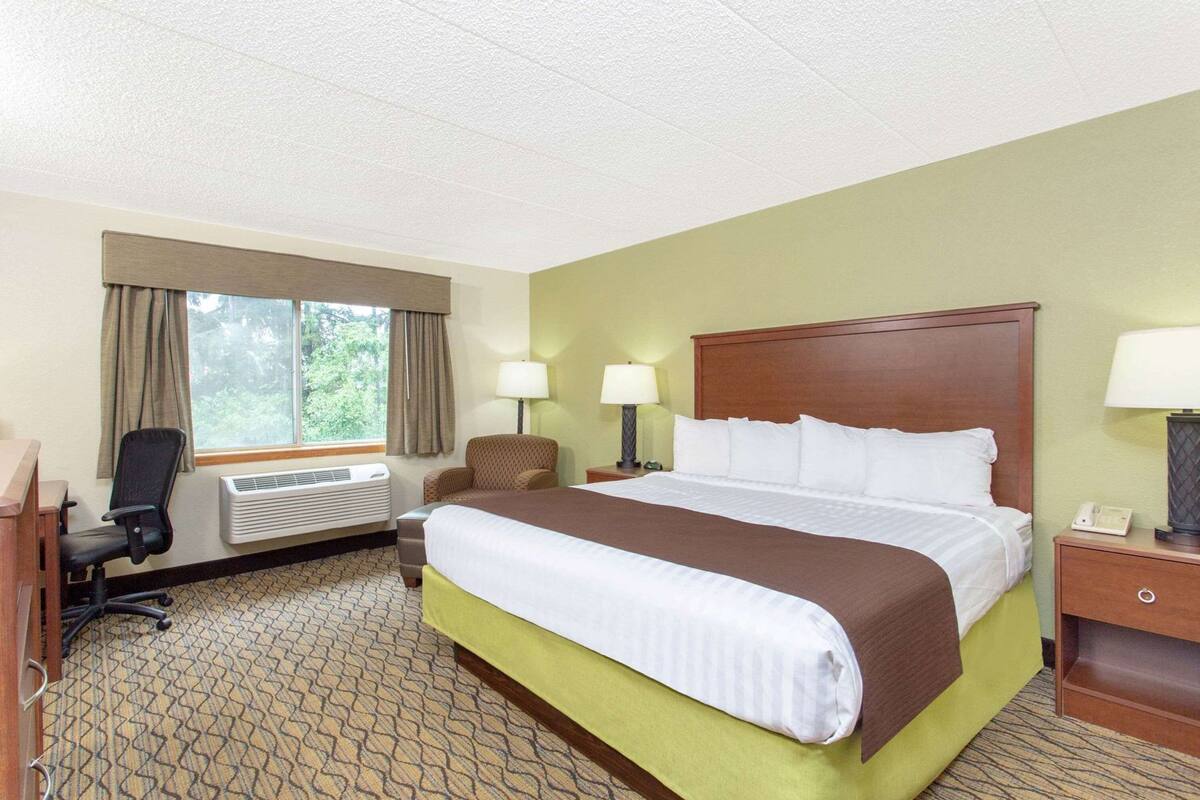 Habitación de hotel con cama, escritorio, silla, ventana con cortinas, unidad de aire acondicionado y mesita de noche con lámpara.