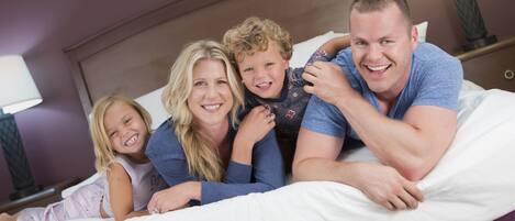 A family of four lying on a bed with white bedding in a hotel room.