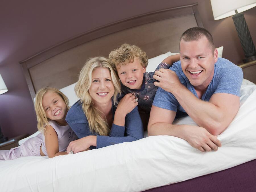 Una familia de cuatro personas acostada en una cama con ropa de cama blanca en una habitación de hotel.