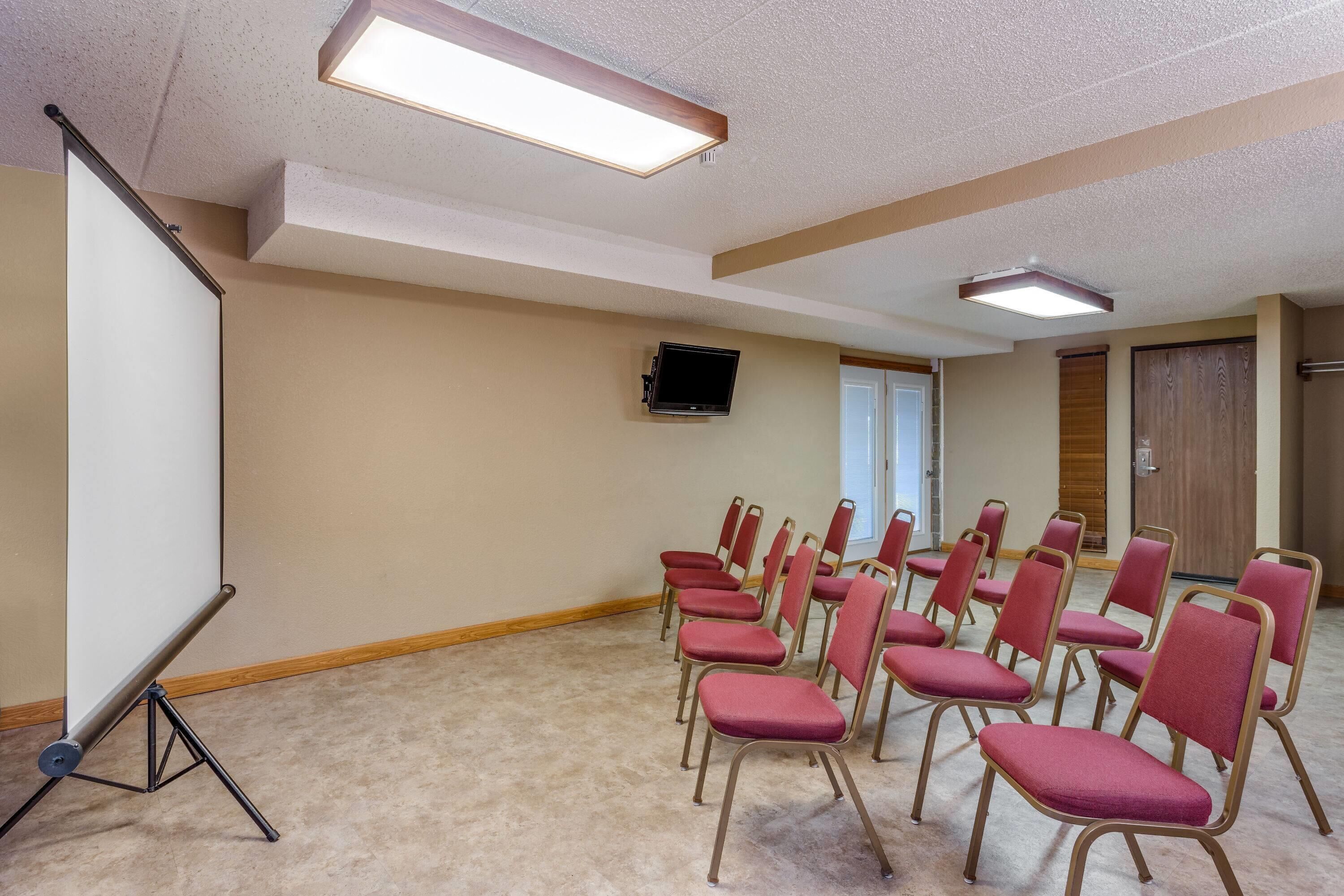 A conference room with beige walls, a projector screen, a flat-screen TV, and rows of red chairs.