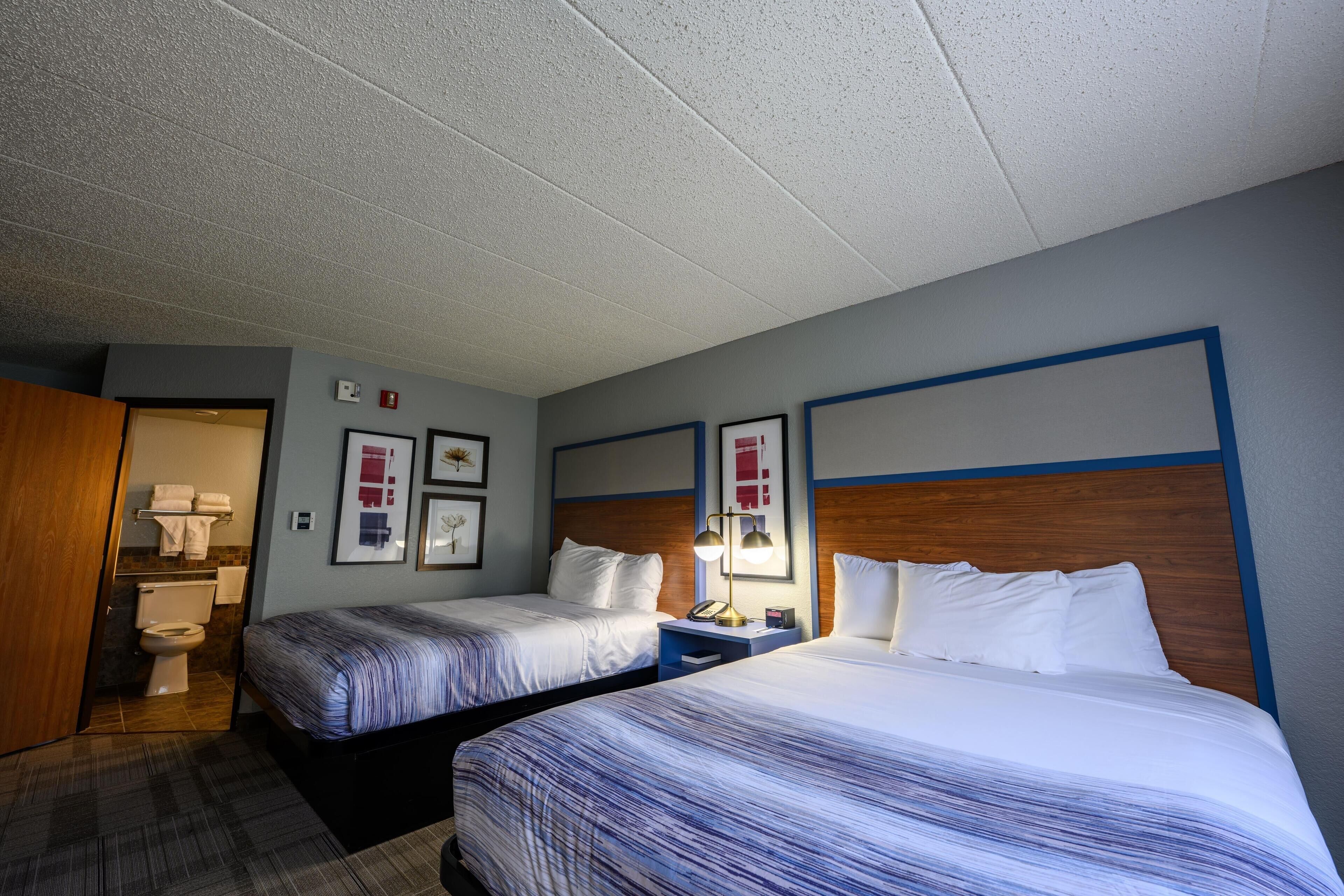 A hotel room with two beds, a bathroom, and a small desk with a lamp.