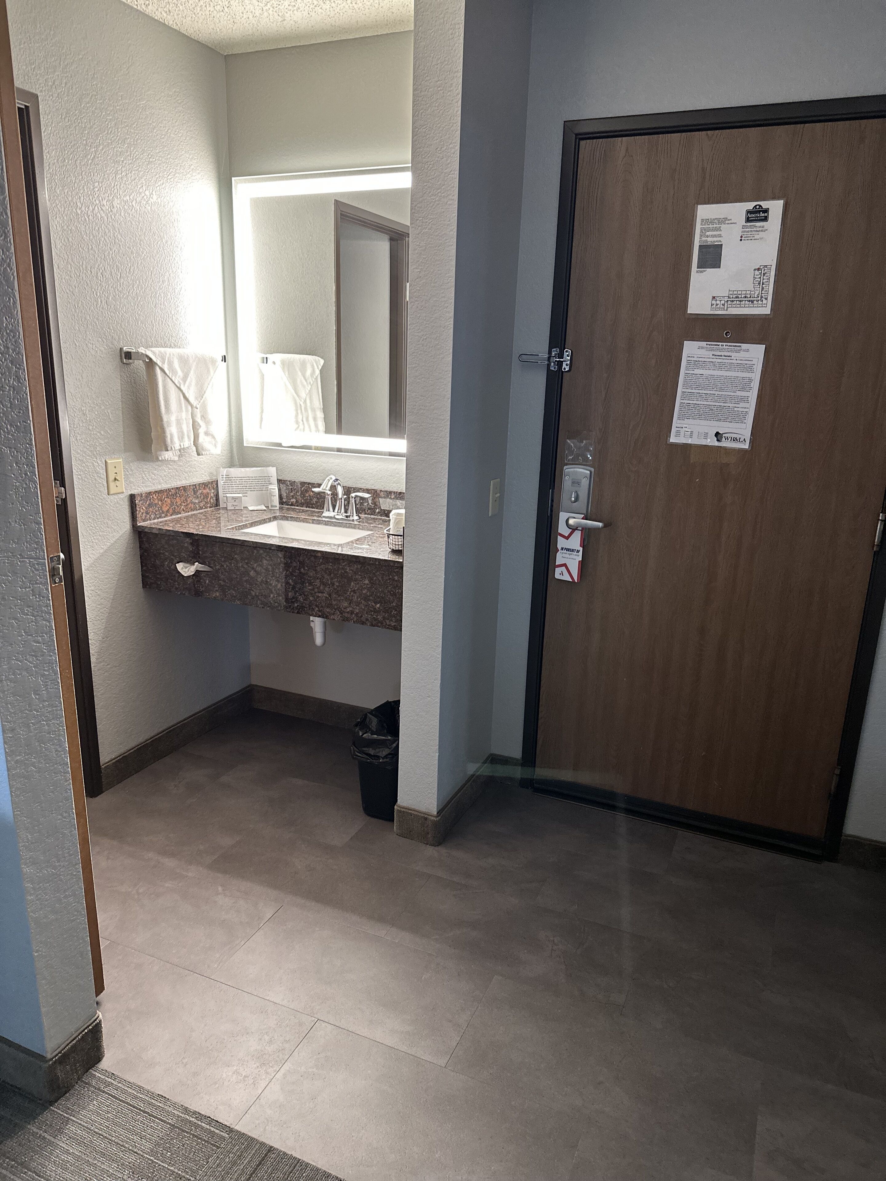 Un baño de hotel con lavamanos de mármol, un espejo, toallero y una puerta con un aviso.