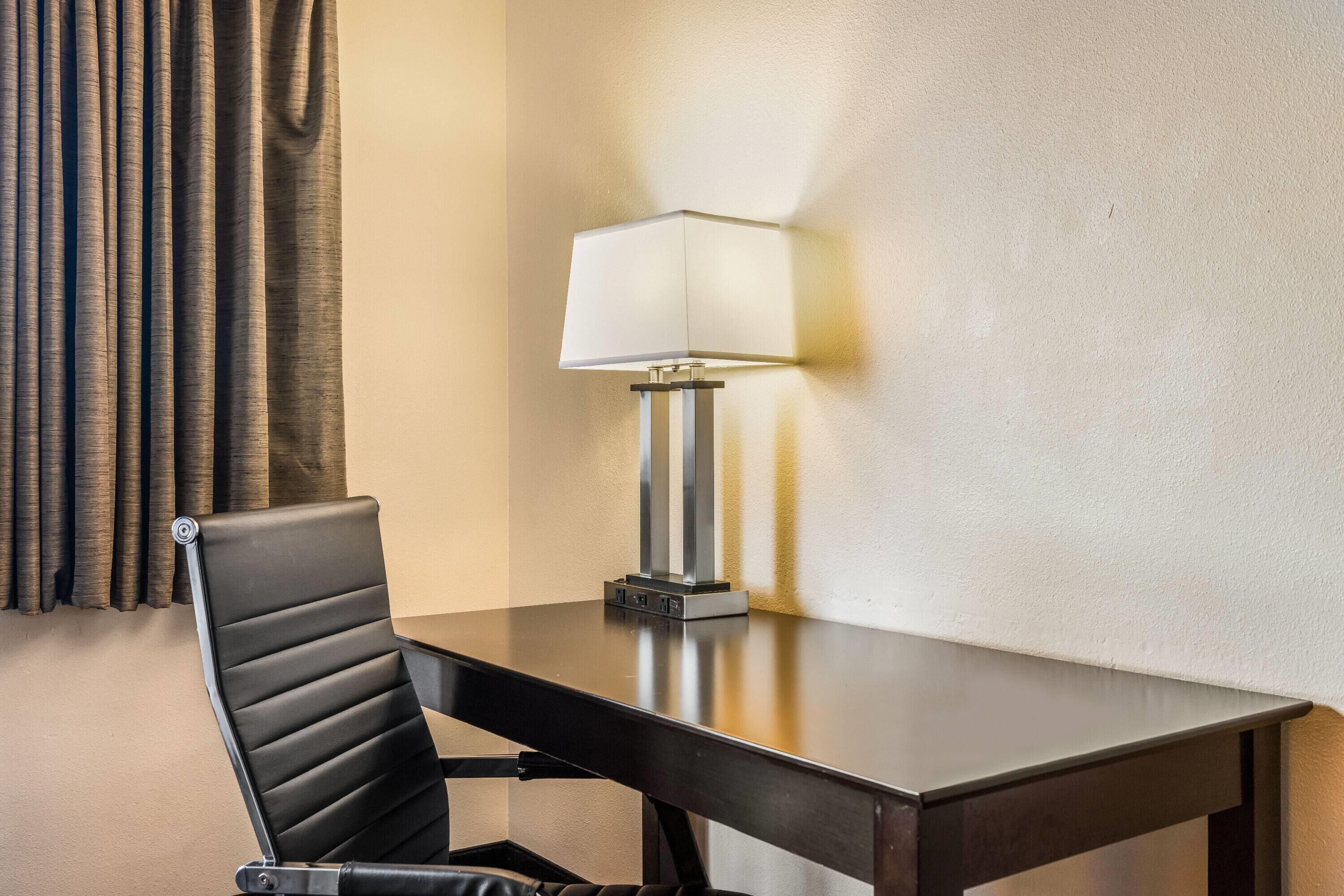 A hotel room with a dark wooden table, a black leather chair, a lamp, and a curtain.