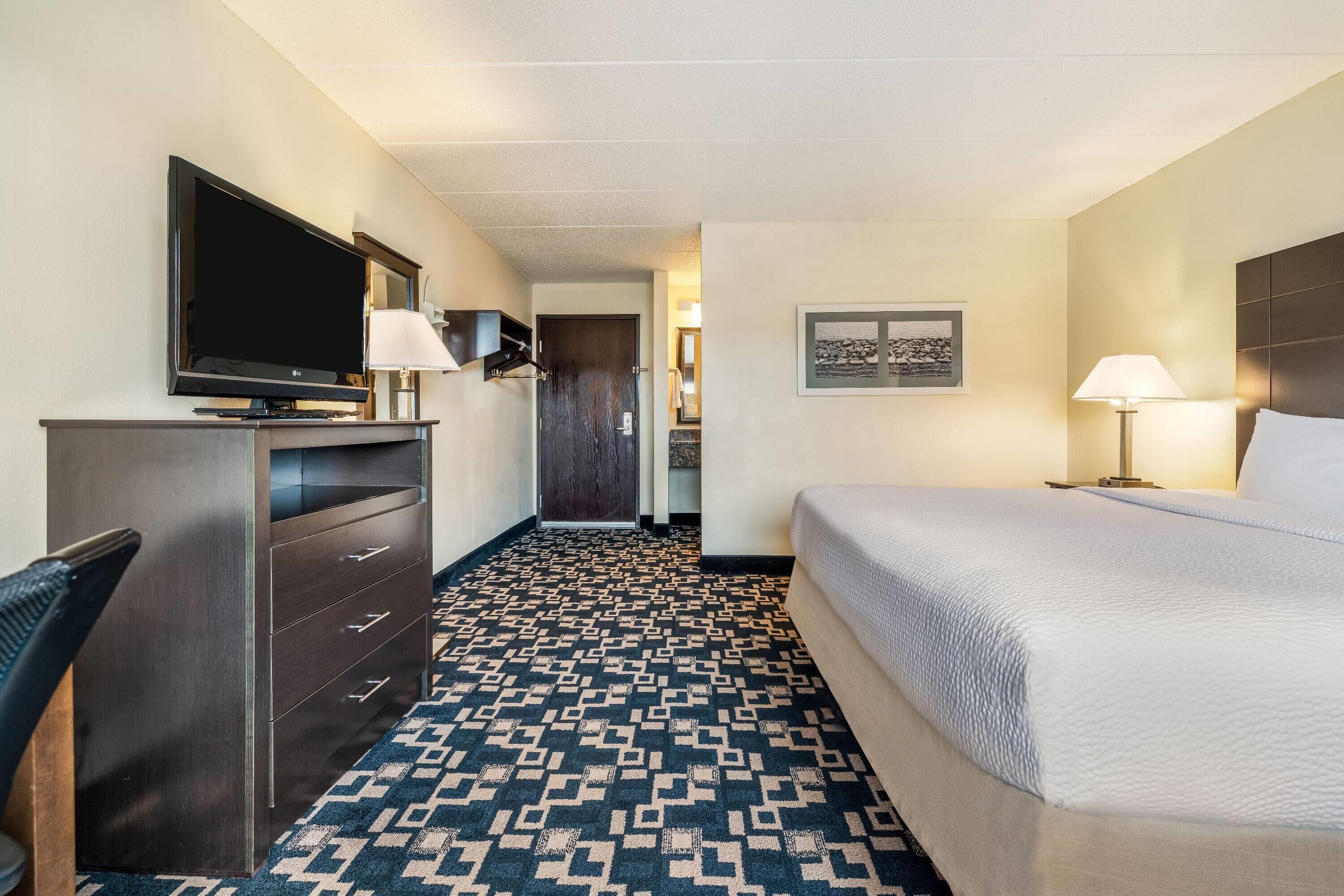 A hotel room with a large bed, a TV on a dresser, a desk with a chair, and a patterned carpet.