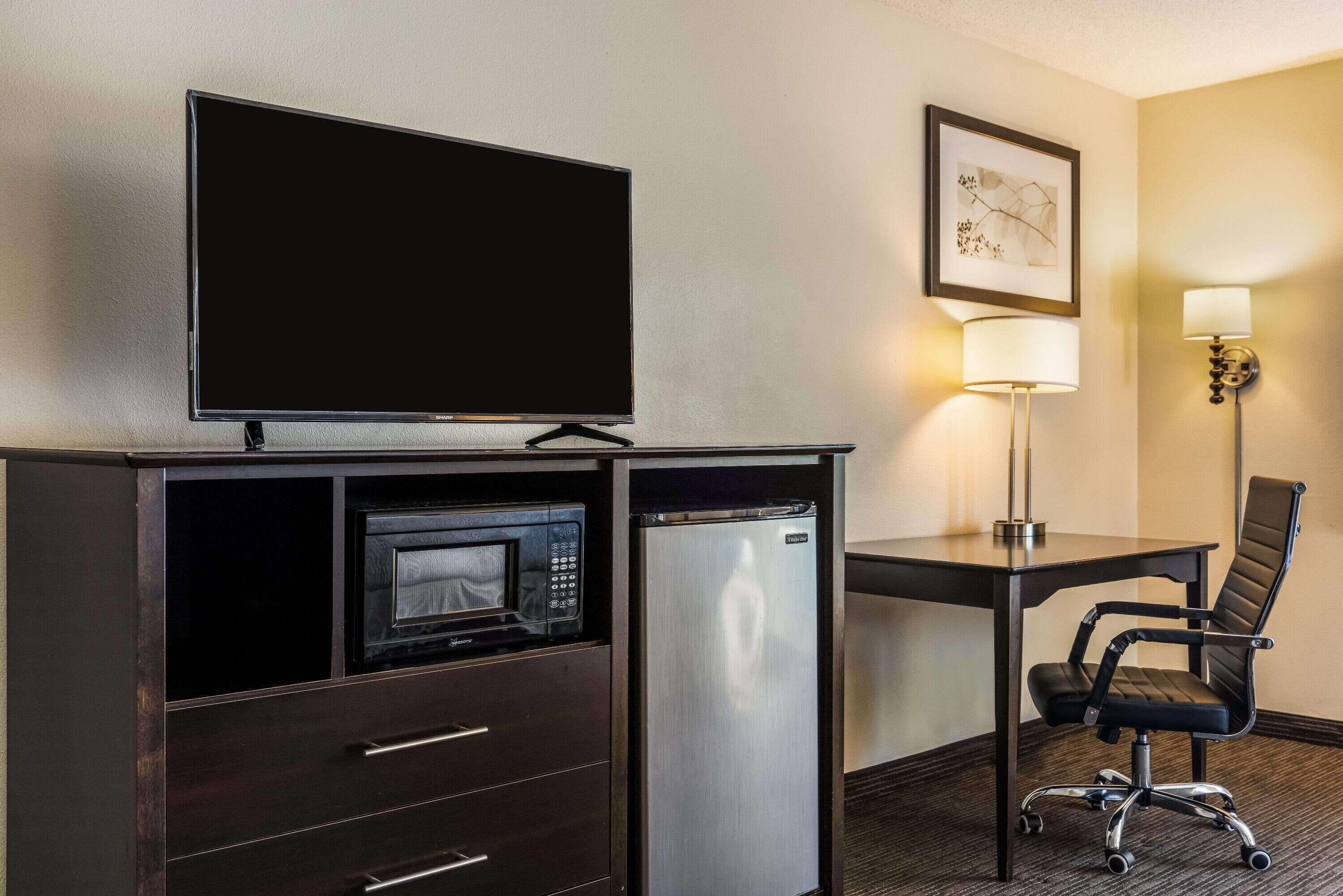 Habitación de hotel con televisión de pantalla plana, microondas, refrigerador pequeño, escritorio con lámpara y una silla.
