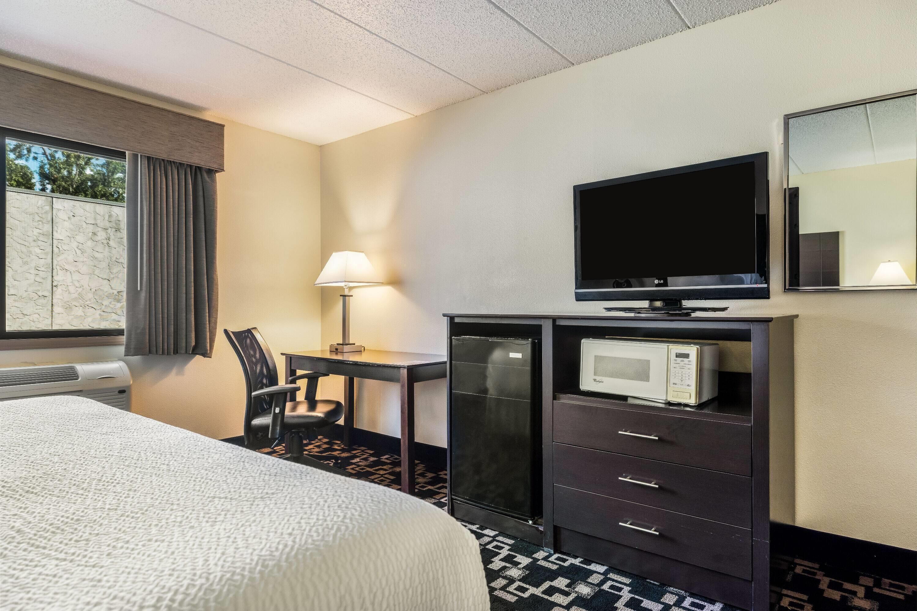 A hotel room with a bed, a desk with a lamp, a TV, a microwave, and a dresser.