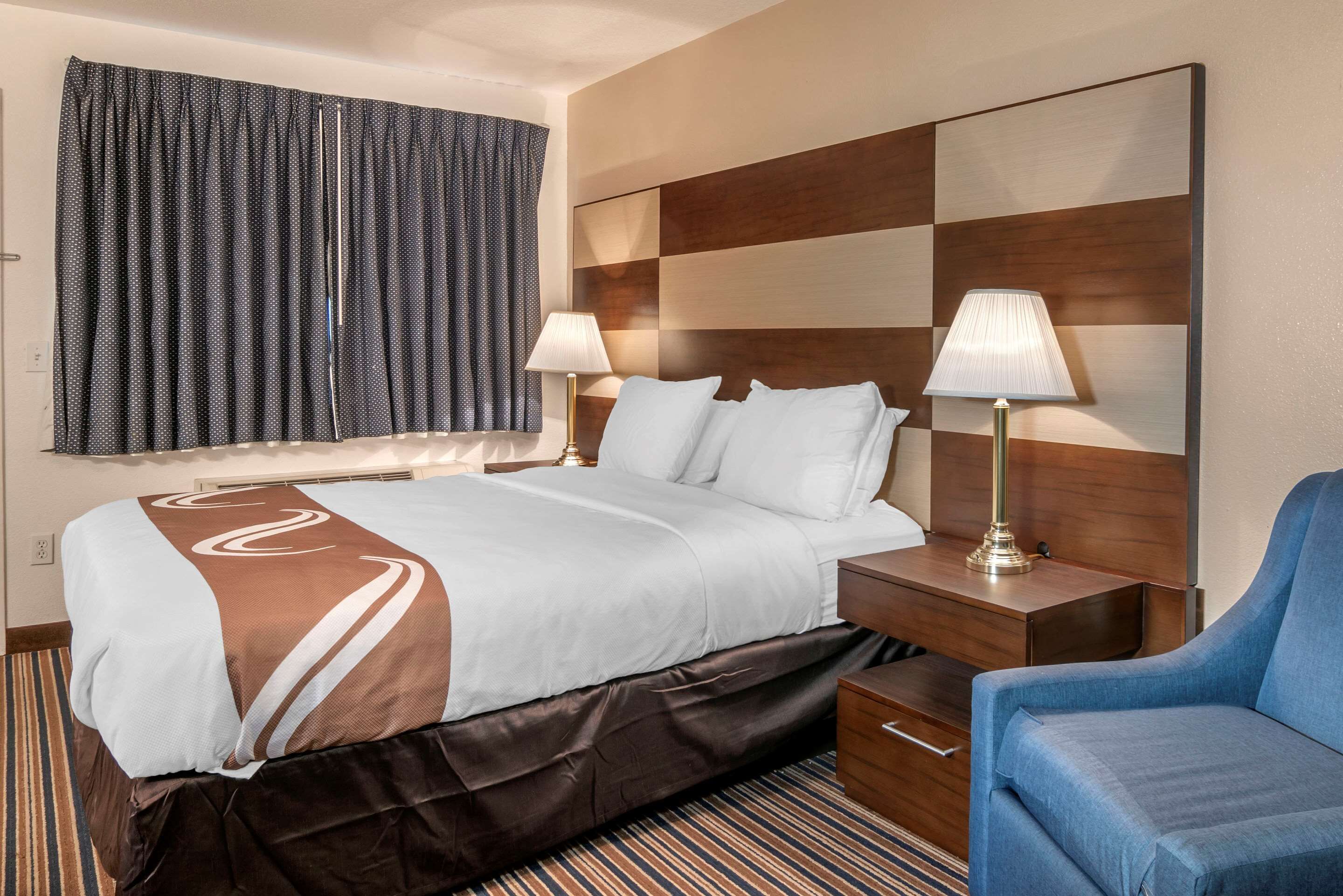 Une chambre d’hôtel avec un grand lit, deux lampes de chevet, une tête de lit en bois et un fauteuil bleu.
