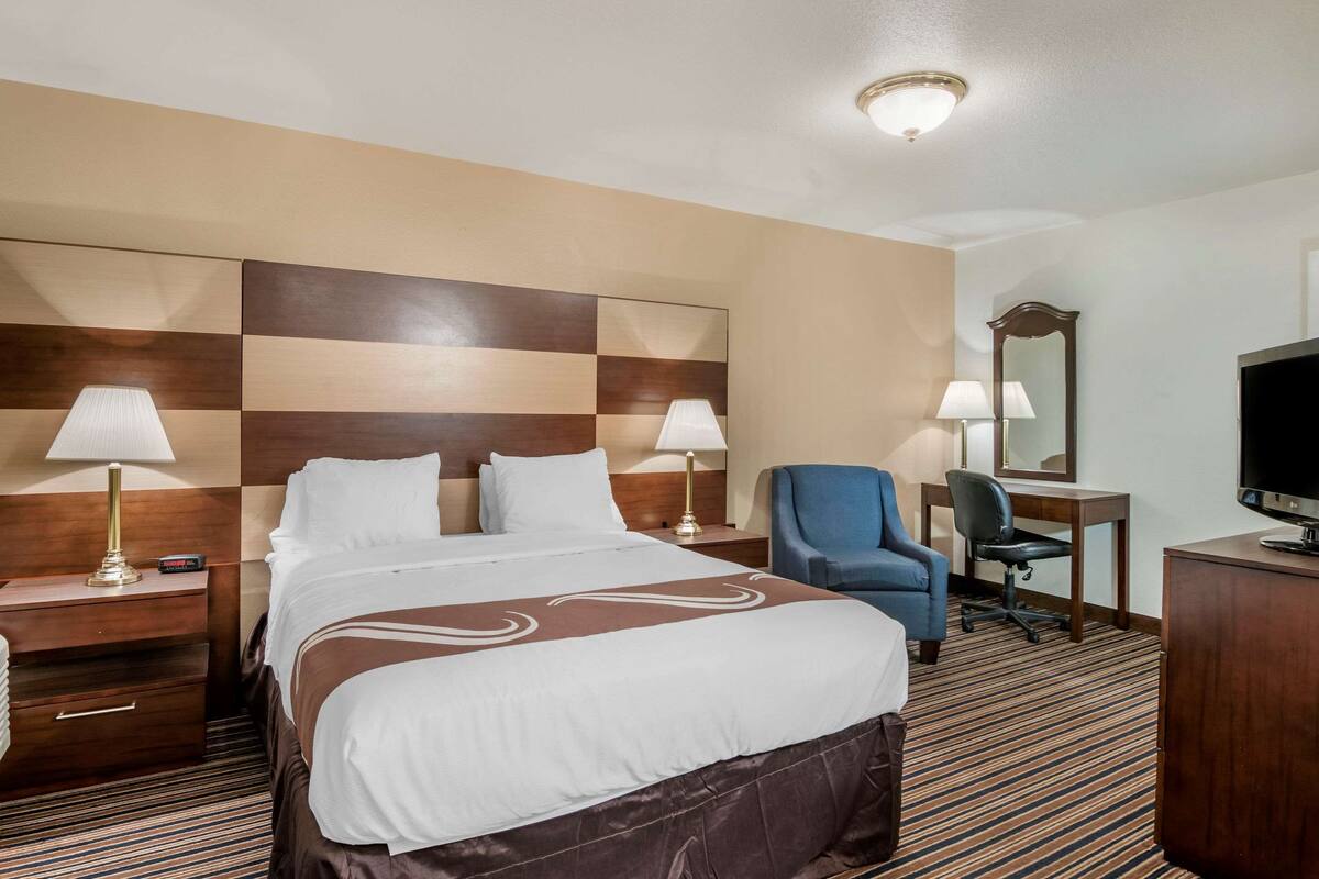A hotel room with a large bed, two bedside tables with lamps, a desk with a chair, a television, and a mirror.