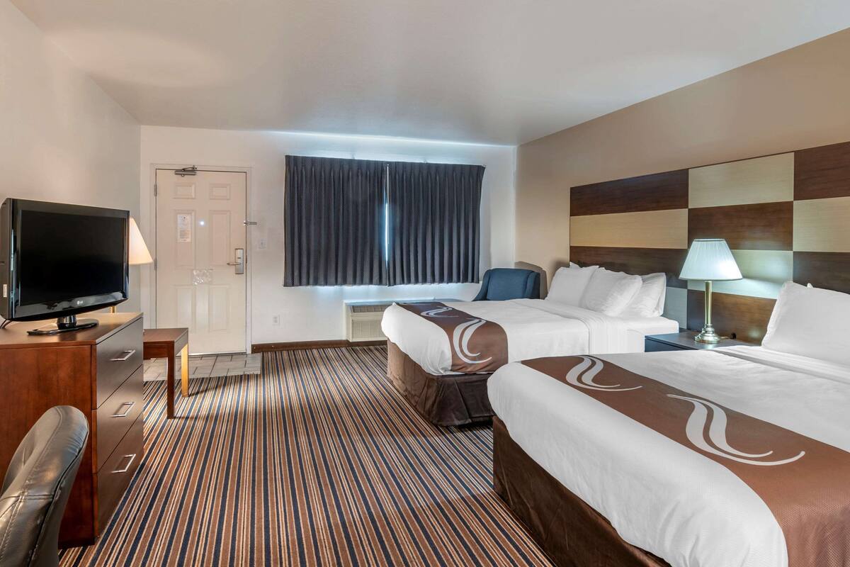 A hotel room with two beds, a TV, a desk, and a large window with curtains.