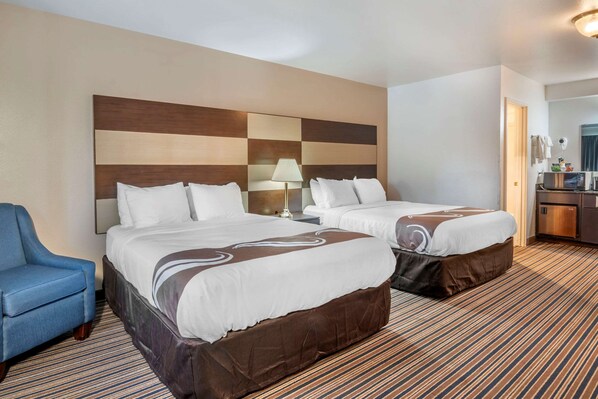 A hotel room with two beds, a blue armchair, a desk with a lamp, and a minibar.