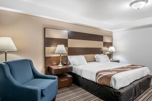 A hotel room with a bed, a chair, a nightstand, a lamp, and a wall clock.