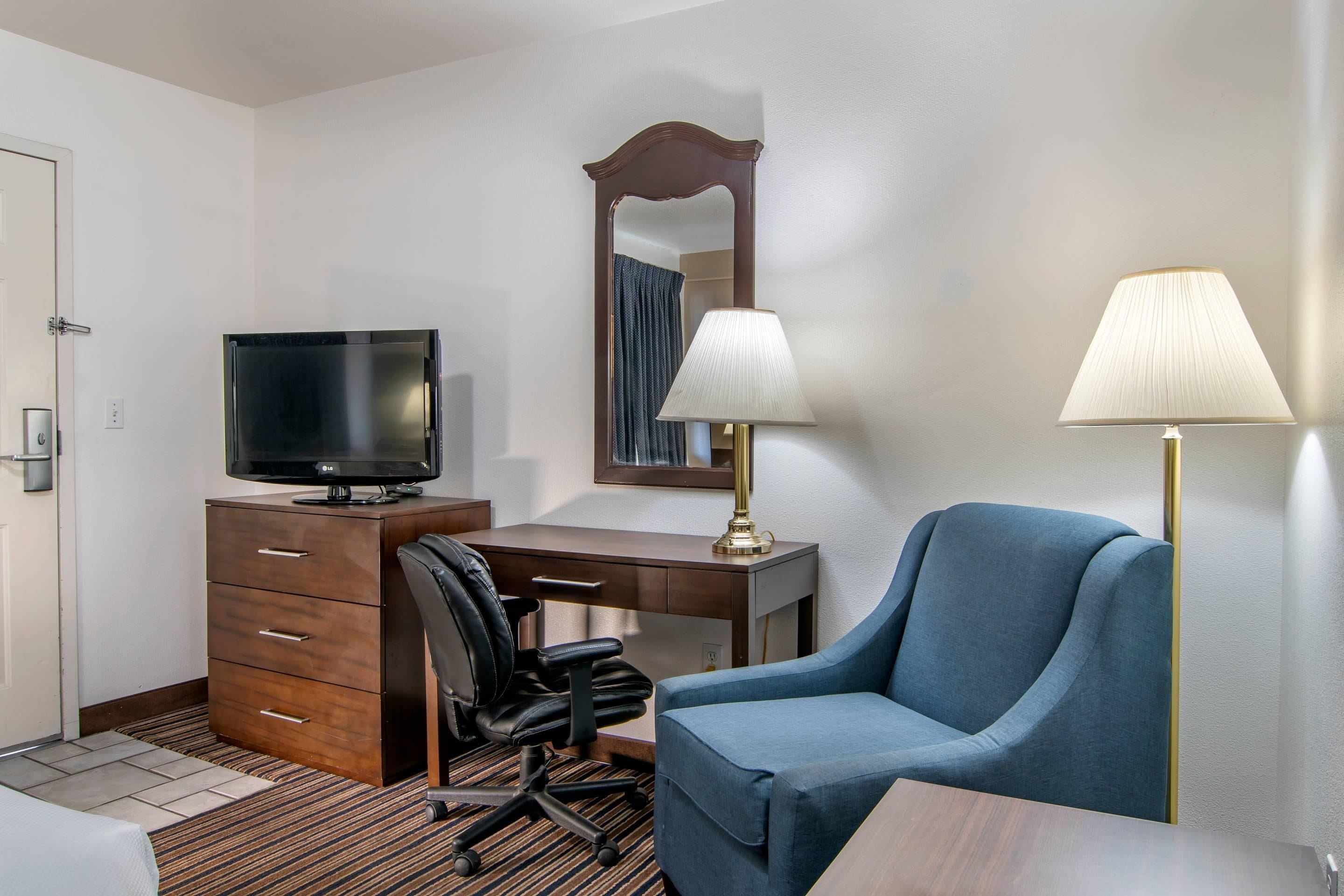 A hotel room with a desk, chair, television, lamp, and a mirror.
