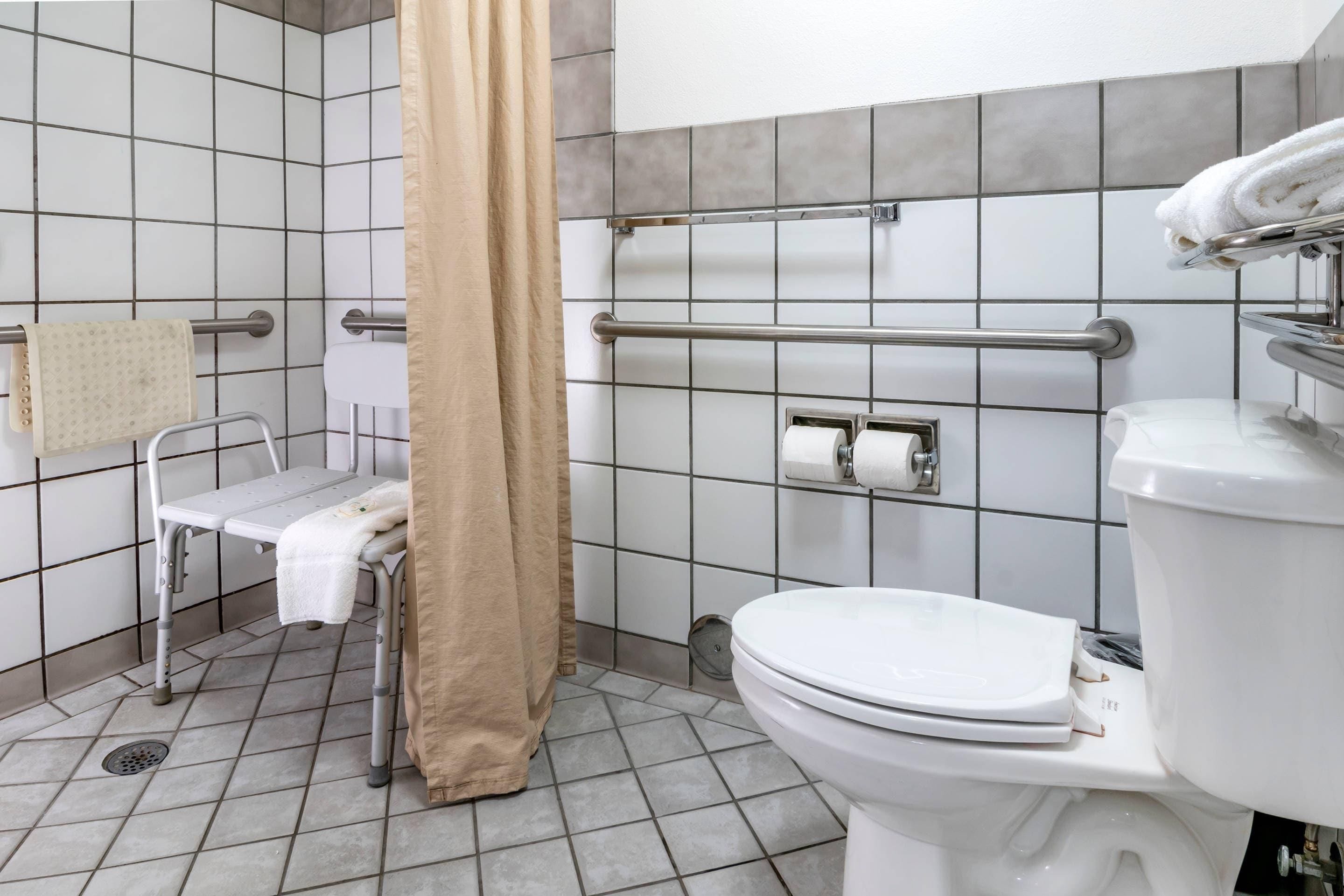 Un baño con inodoro blanco, ducha con cortina color beige, barra de apoyo, toallero y un lavamanos pequeño y blanco.