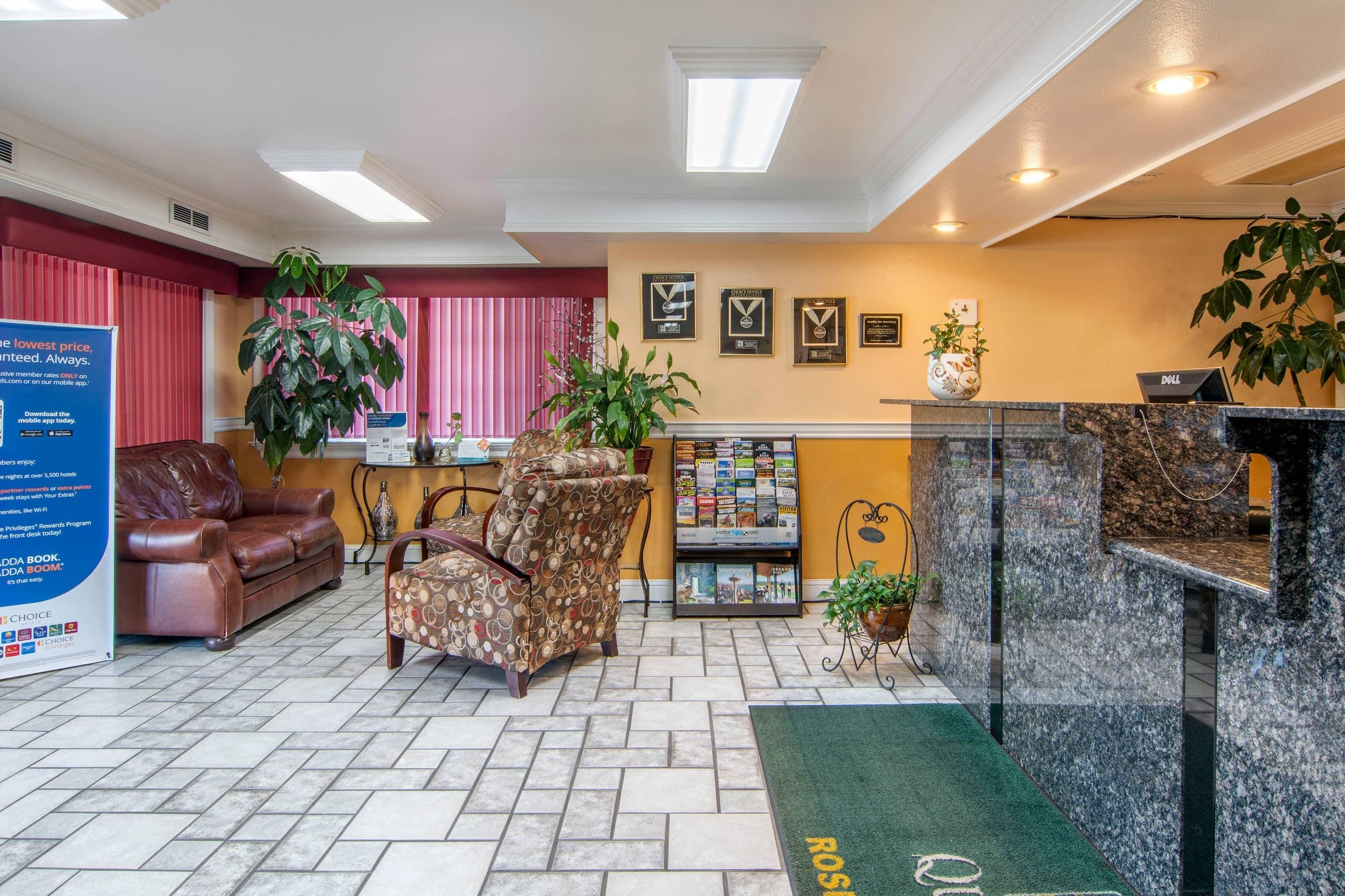 Un hall de hotel con mostrador de recepción, una zona de estar con sofá y sillón, una planta grande y un tablón de anuncios con premios enmarcados.