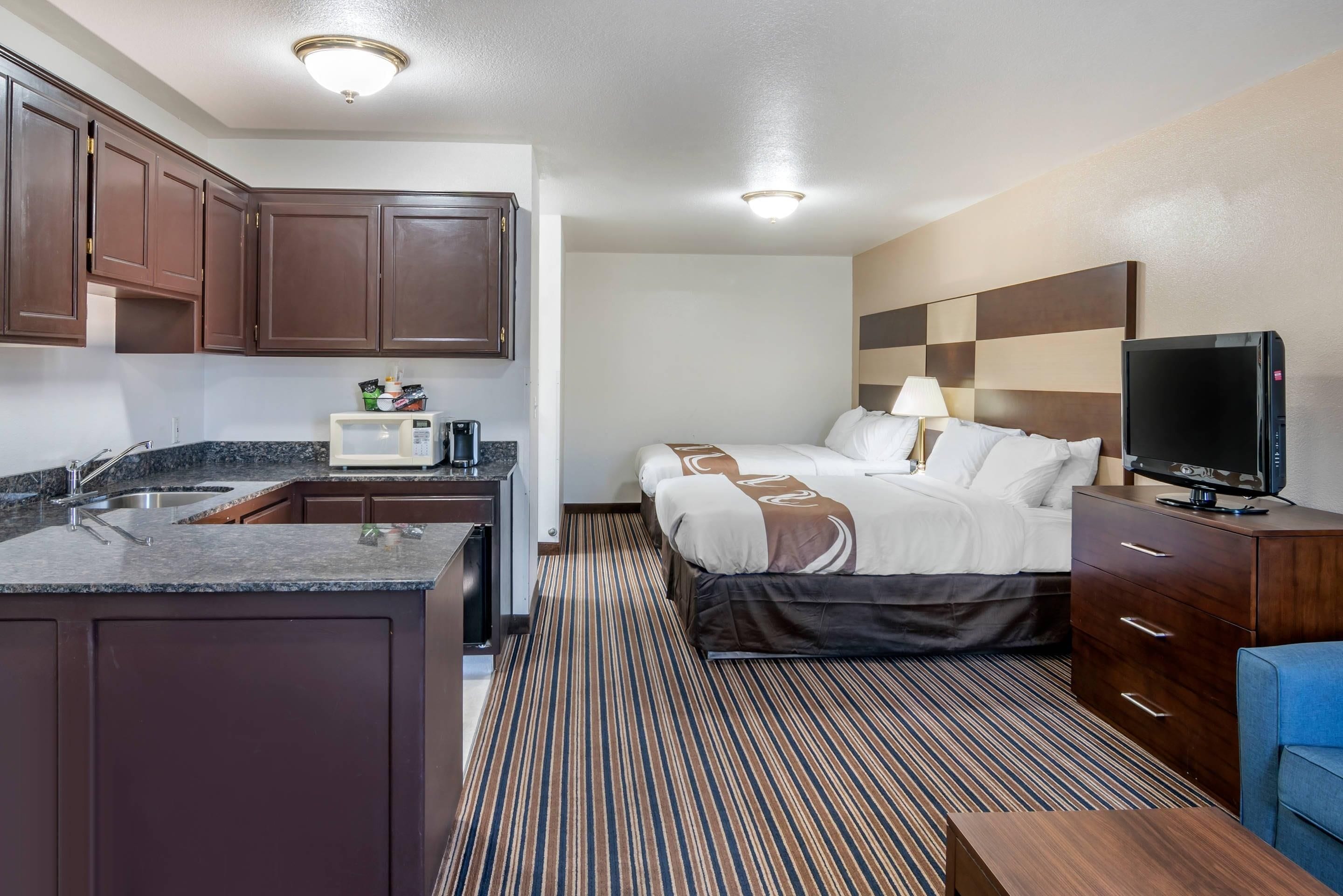 Une chambre d’hôtel avec une kitchenette, un lit recouvert d’une courtepointe à rayures, une télévision et un fauteuil bleu.